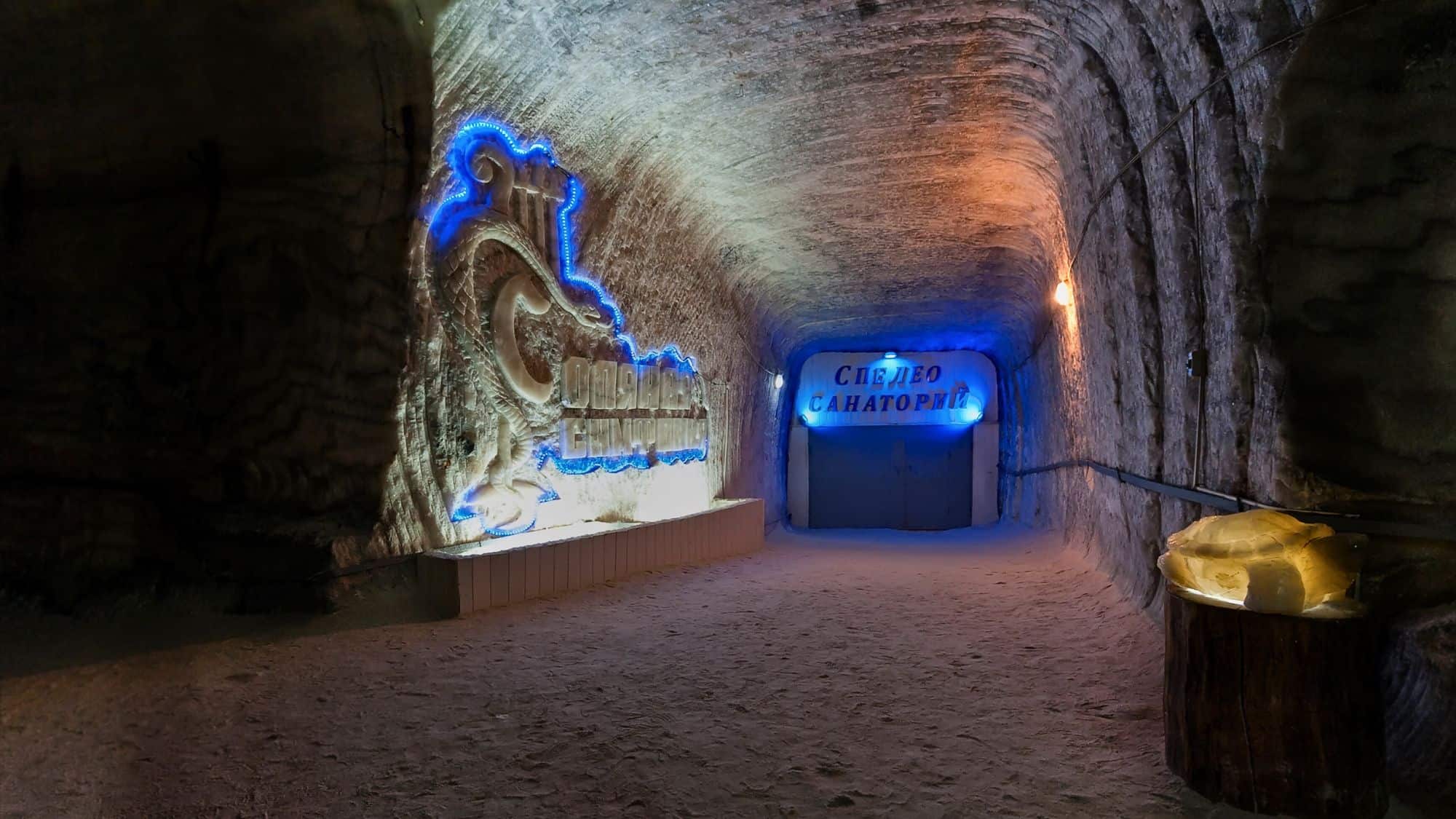 A softly lit hallway inside a salt mine with Russian signage and a glowing dragon mural marks the entrance to a speleo-sanatorium.