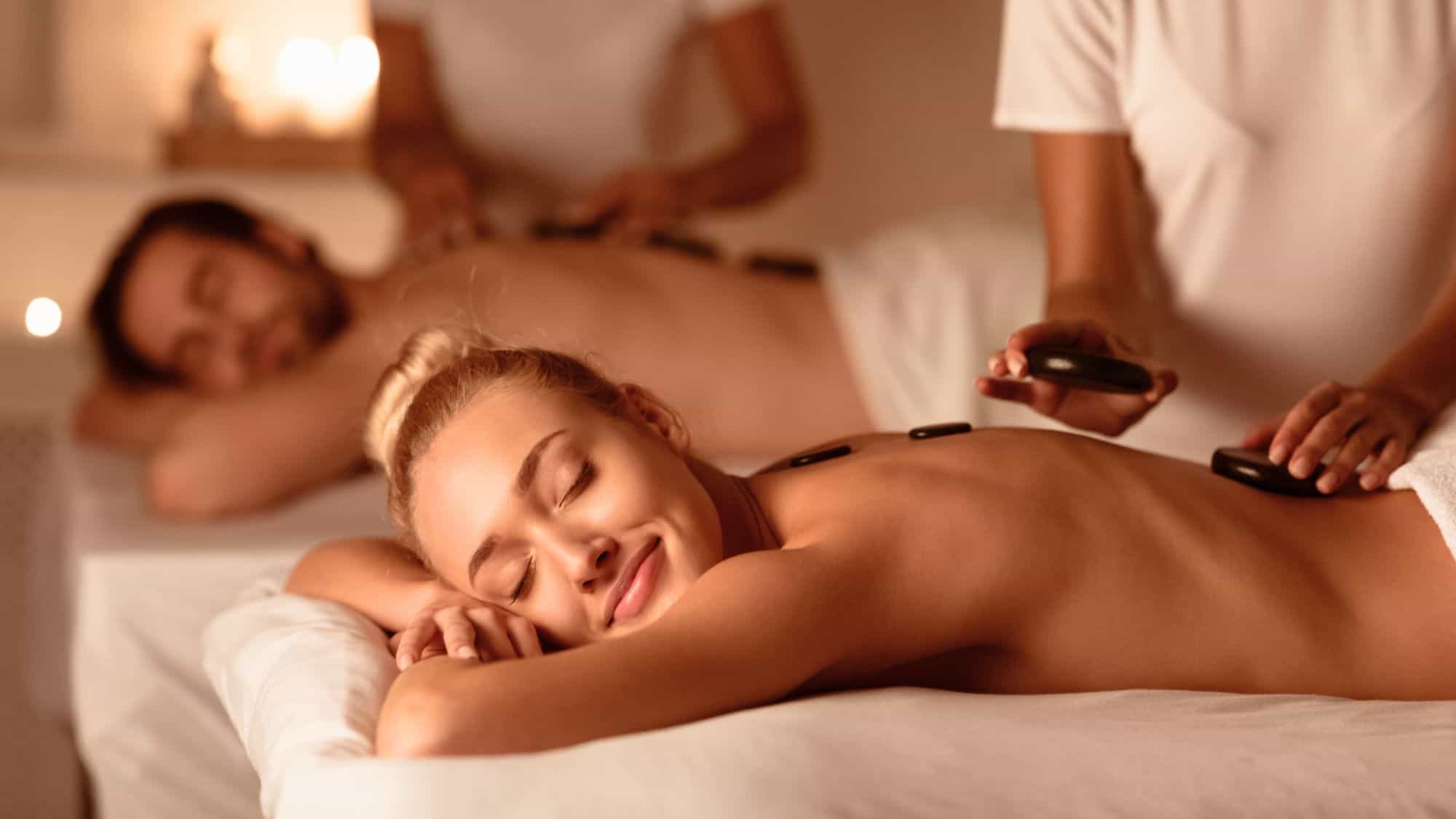 A smiling woman and a man enjoy a relaxing hot stone massage side-by-side in a candlelit cruise ship spa setting.