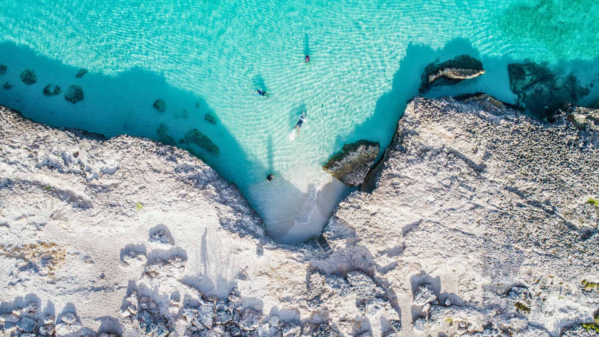 A tranquil coastal area with clear turquoise water and a rocky beach. A few people are seen enjoying the shallow waters, highlighting the location's relaxing vibe.