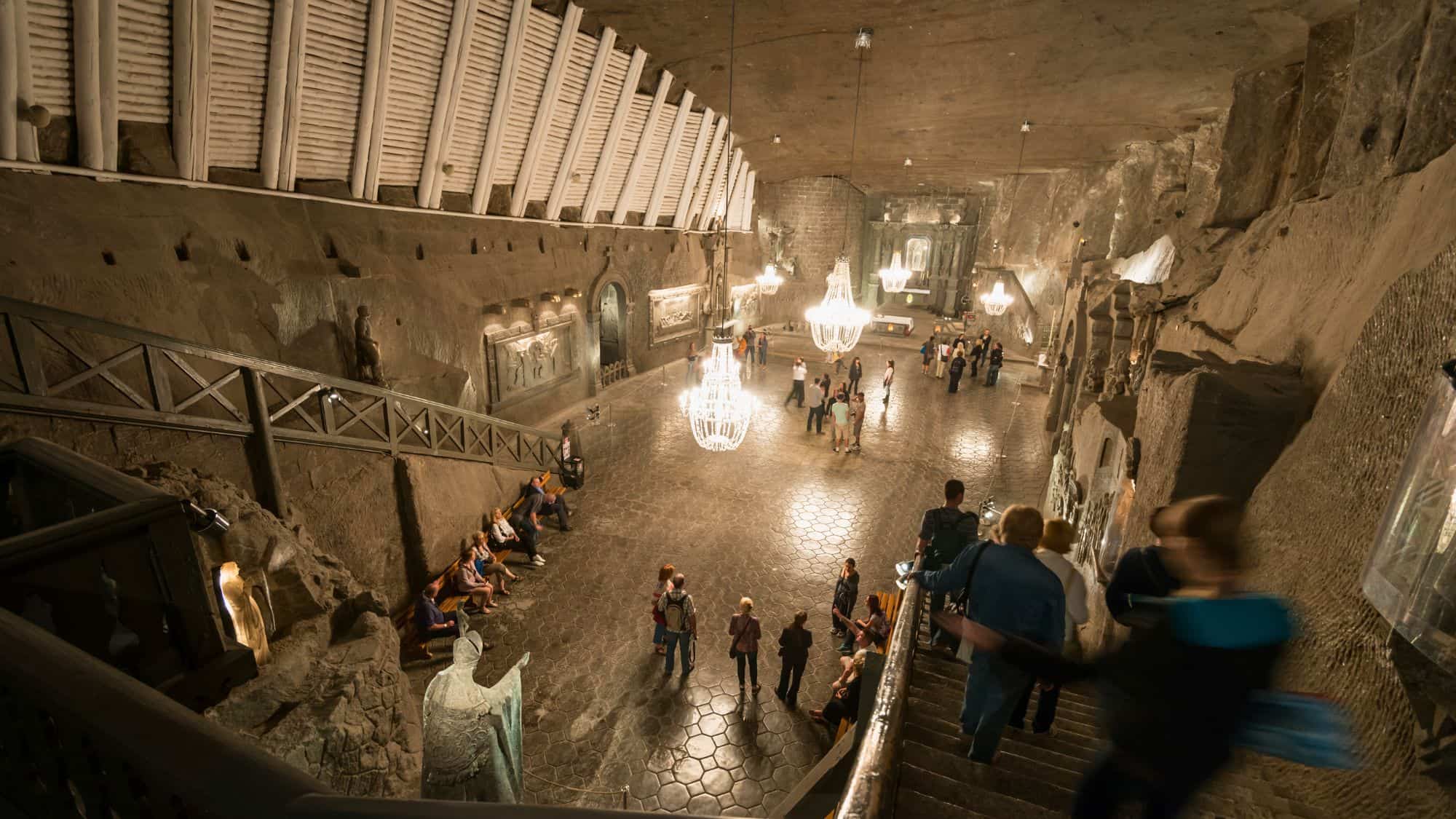 A grand subterranean chapel carved from salt, complete with chandeliers, religious sculptures, and visitors exploring the vast hall.