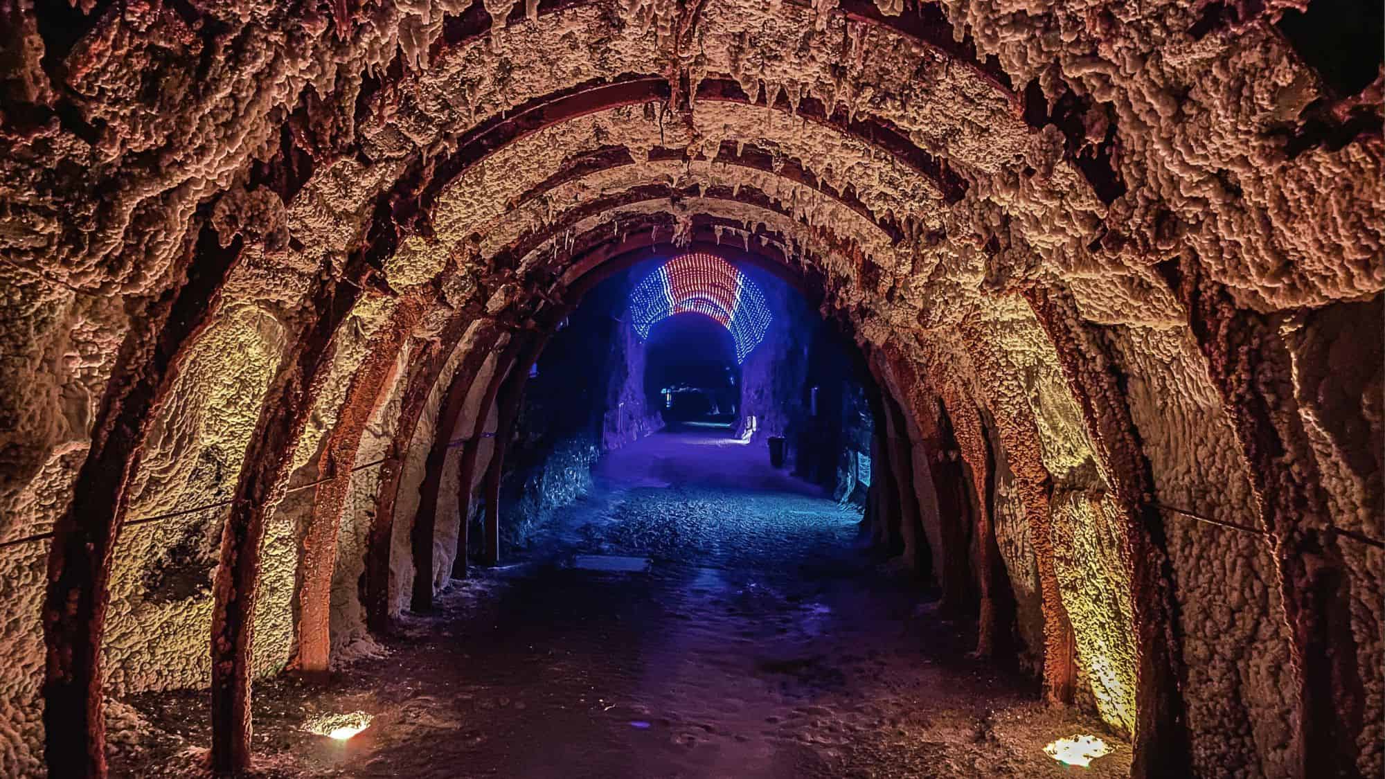 A narrow salt-covered tunnel with arched metal supports glows with amber and blue lighting, leading to a vibrant LED display in the distance.