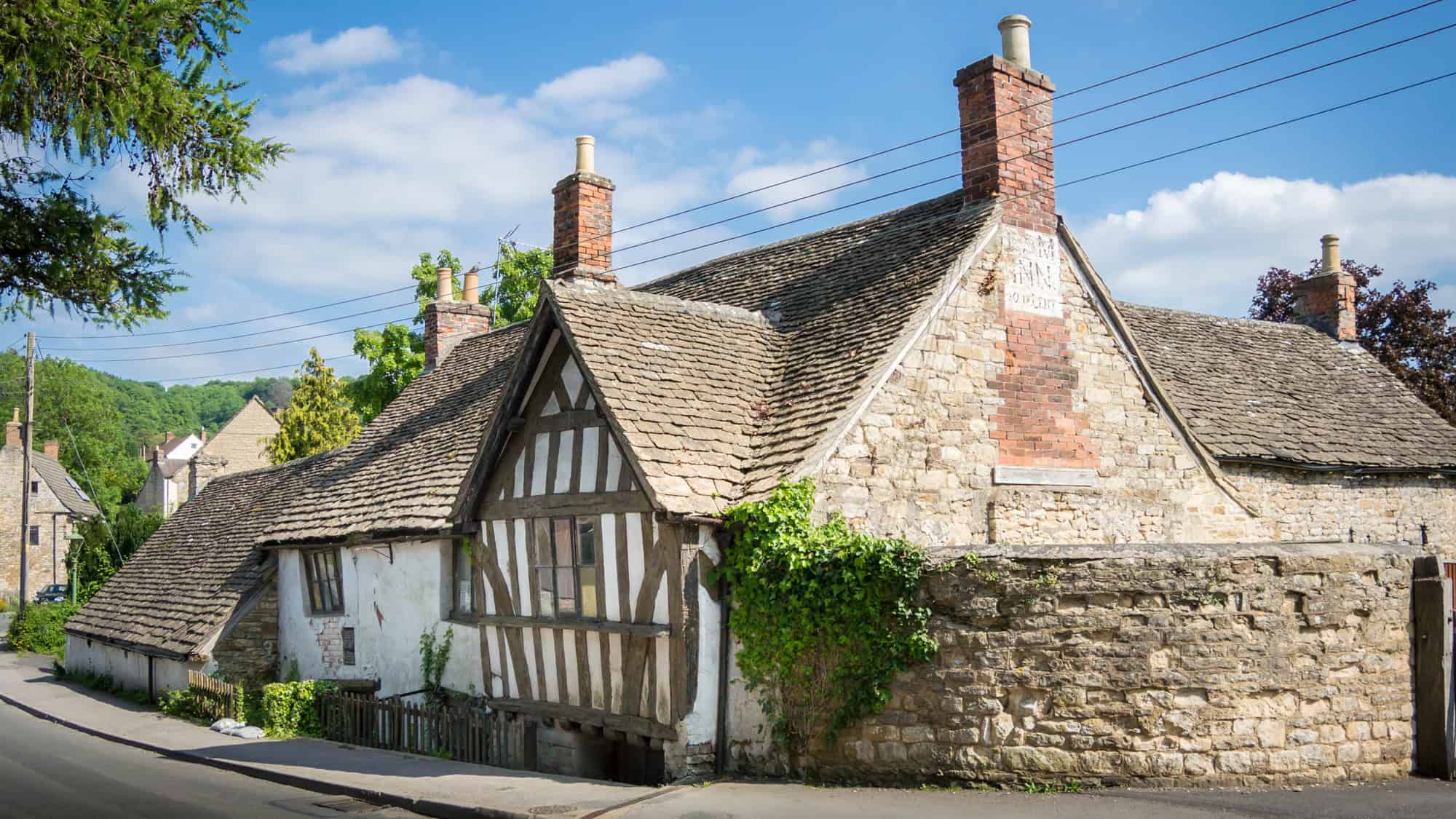 Charming old stone cottage in the Cotswolds with a steep, tiled roof, timber-framed gable, and ivy climbing up the rustic walls under a blue sky.
