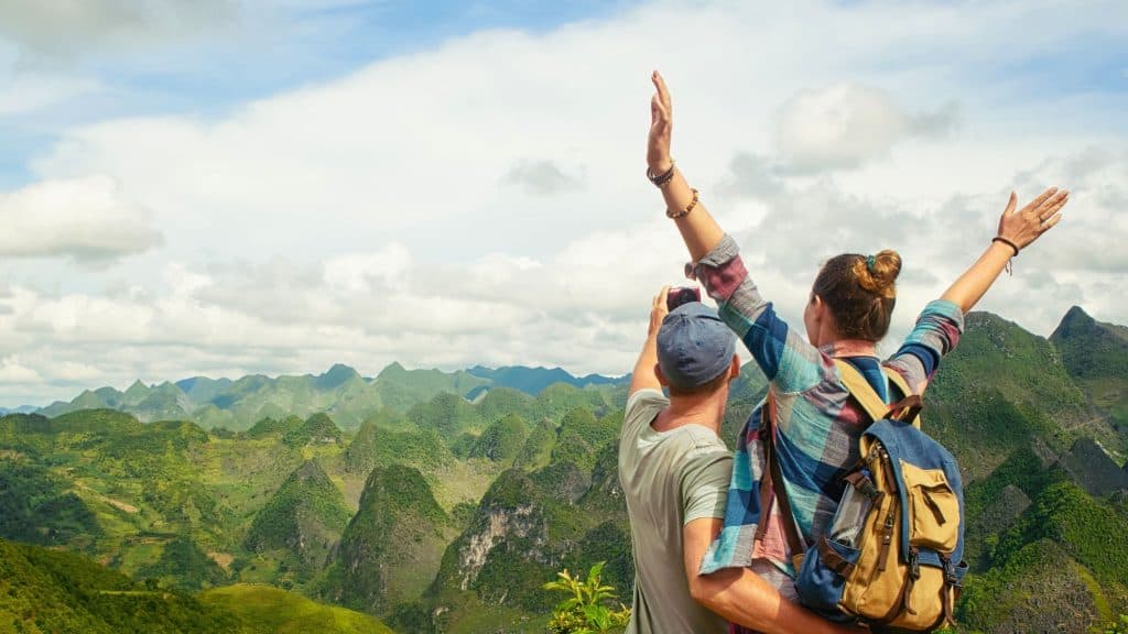 Two individuals enjoying a panoramic view of lush, green mountains under a partly cloudy sky. One is taking a photo, while the other raises their arms in excitement, emphasizing adventure and exploration.