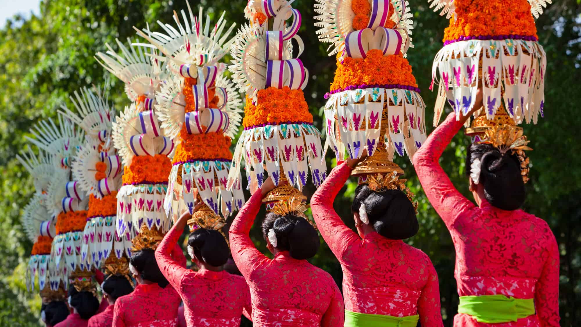 Women in matching pink kebaya with green sashes carry tall, elaborate ceremonial offerings made of flowers and palm leaves on their heads during a traditional Balinese procession.