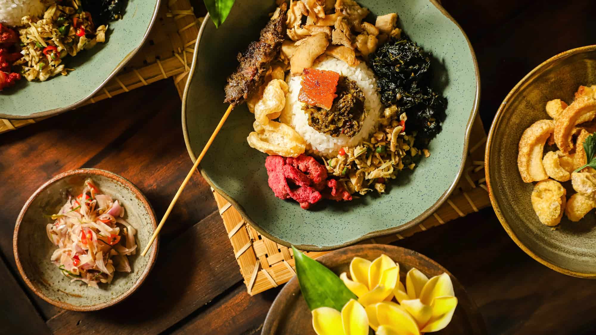 A beautifully plated Balinese dish featuring a mound of white rice surrounded by assorted meats, vegetables, crispy crackers, a satay skewer, and sambal, served on traditional earthenware.