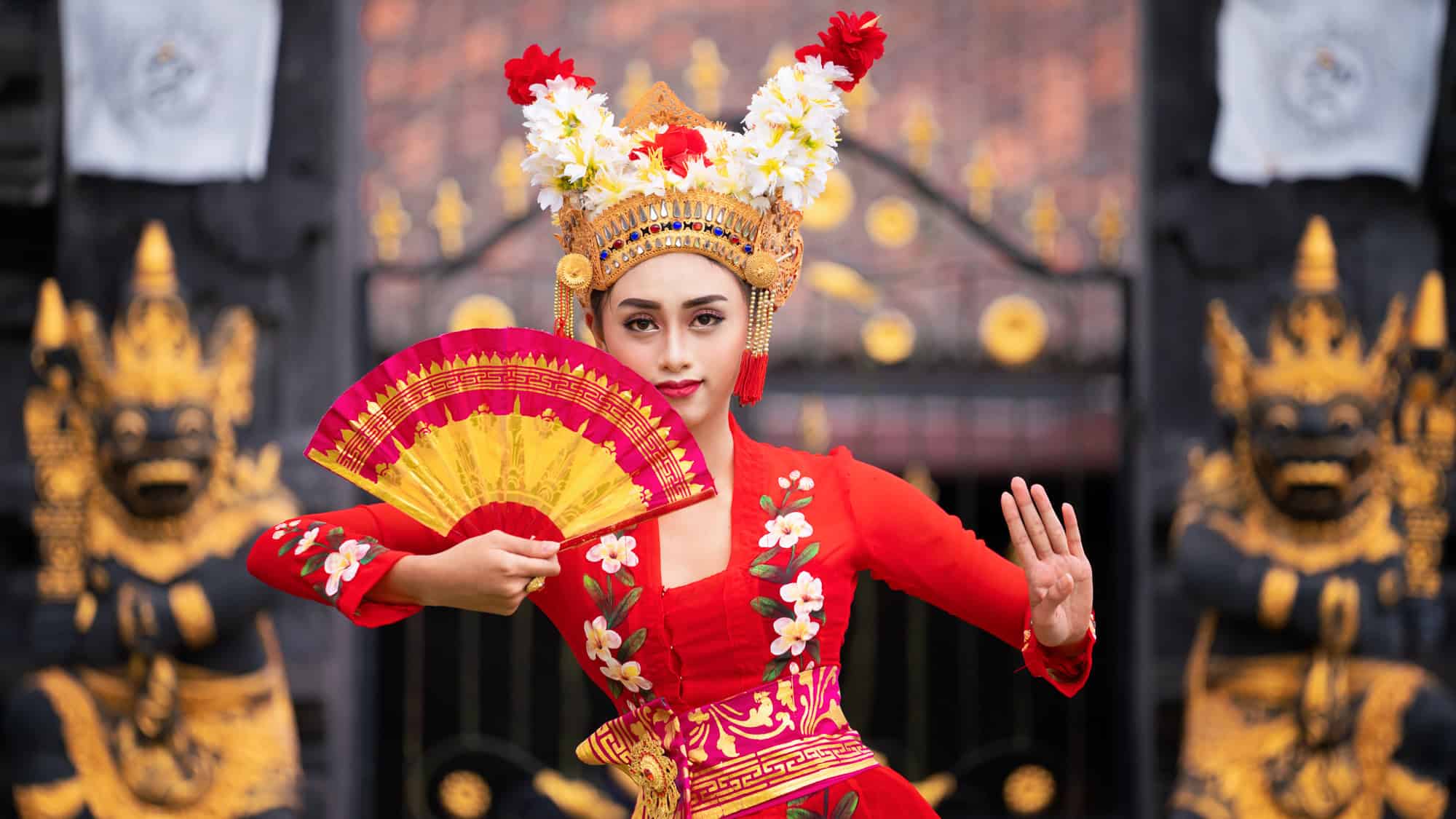 A Balinese dancer dressed in a vibrant red outfit with floral embroidery poses mid-performance, holding a red and yellow fan, wearing an ornate gold headdress adorned with flowers, in front of a traditional temple gate.