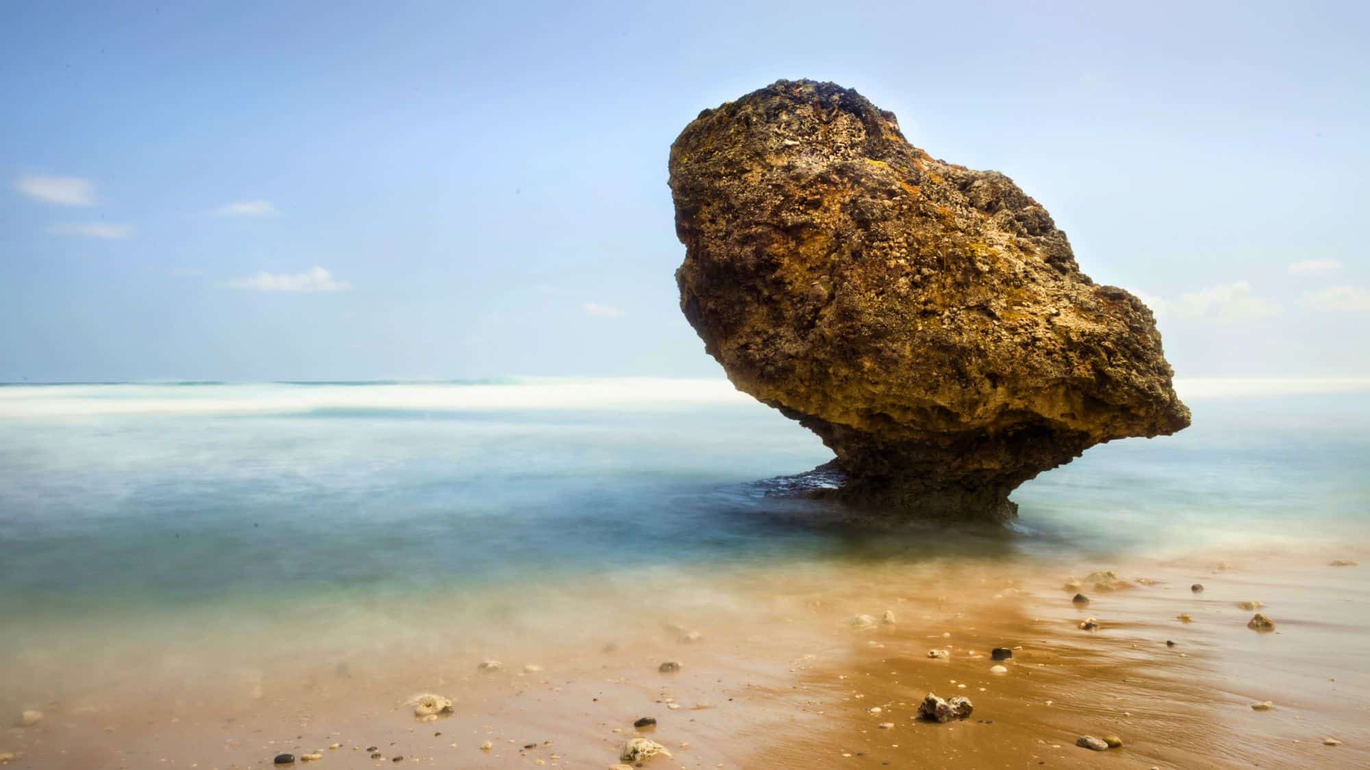 A large, mushroom-shaped coastal rock stands on wet sand surrounded by soft, misty waves under a pastel blue sky.