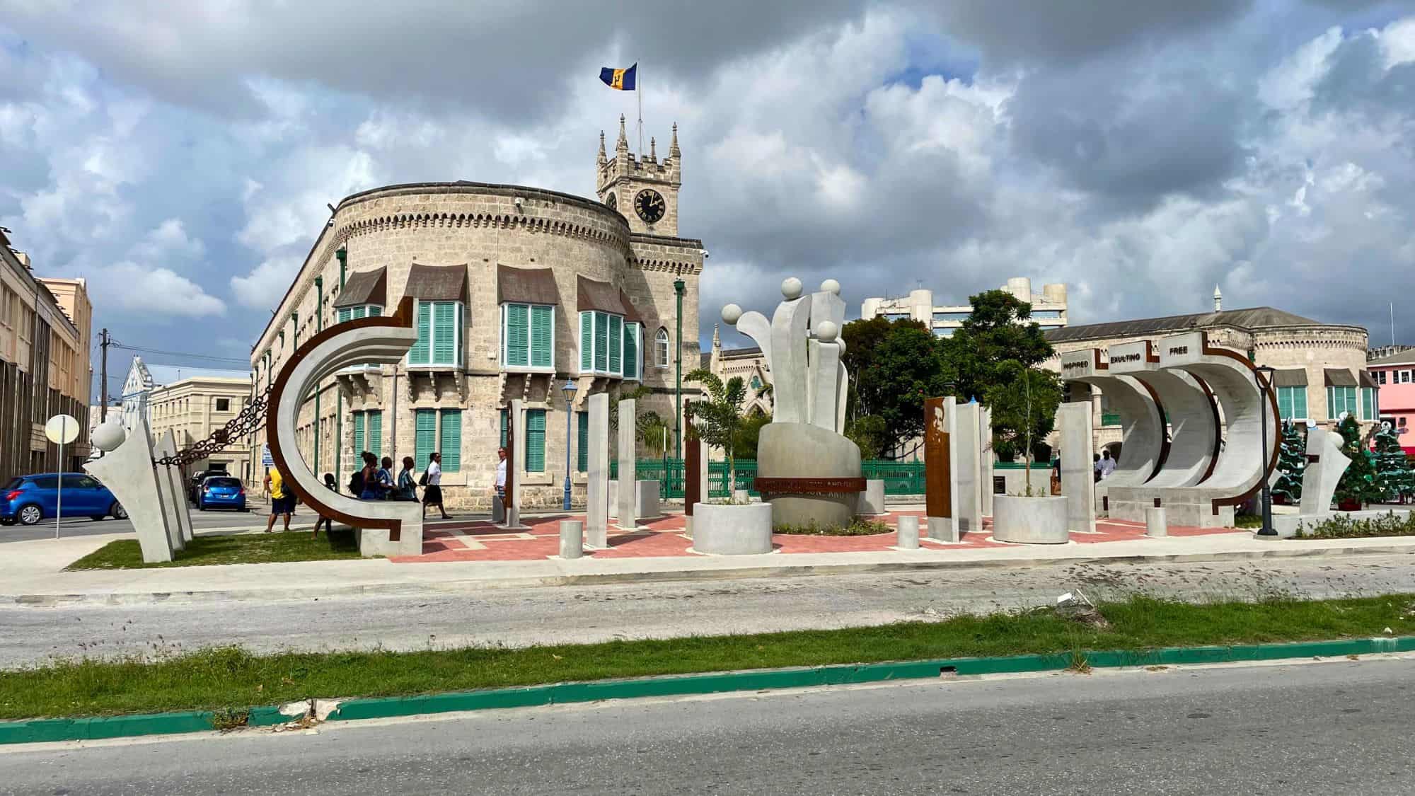 Modern concrete sculpture commemorating emancipation stands in front of the Parliament Buildings in Bridgetown, with the Barbados flag flying atop a clock tower.