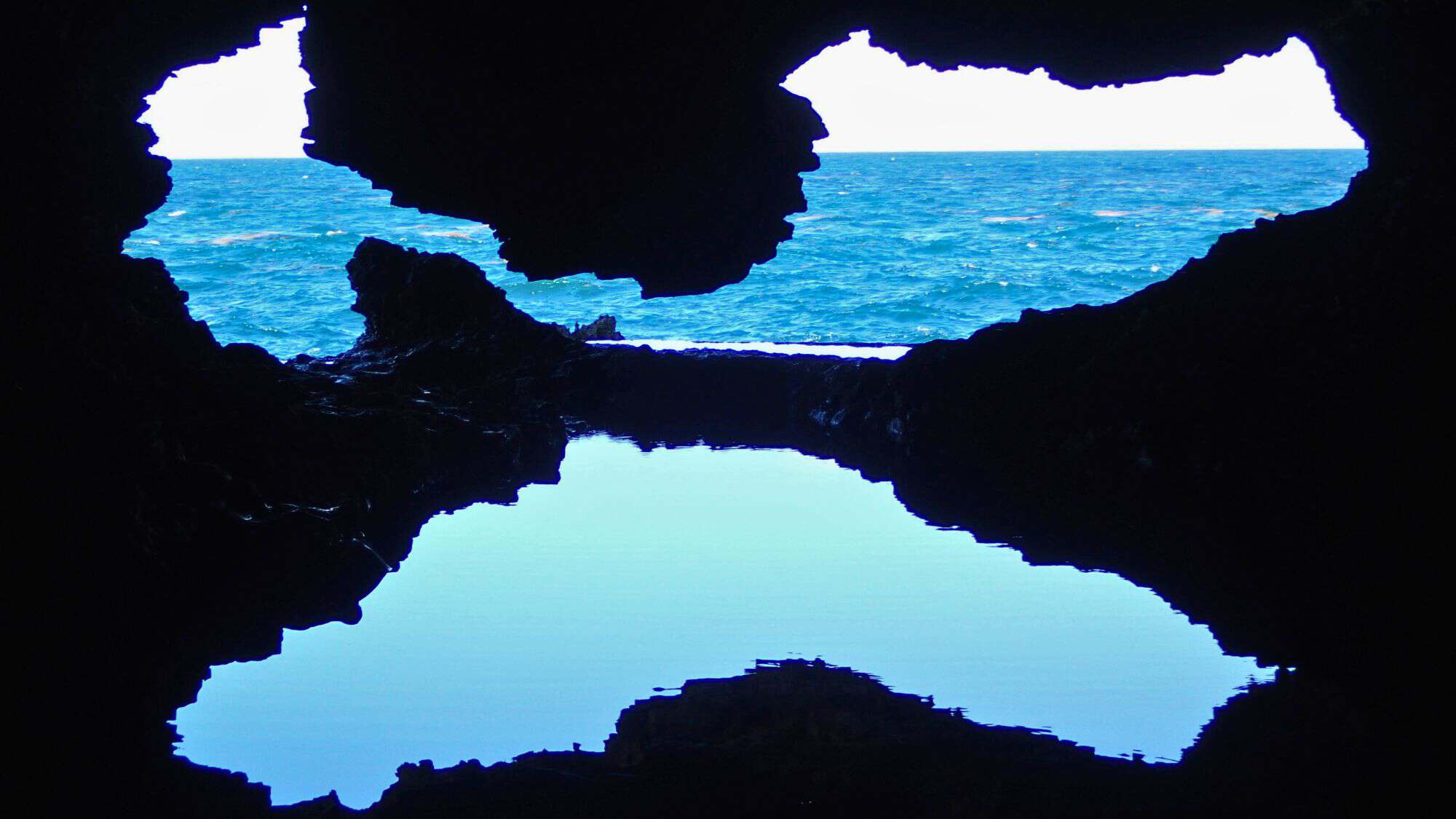 A view of the ocean through the jagged black rock openings of Animal Flower Cave in Barbados, with calm blue water reflecting the sea beyond.