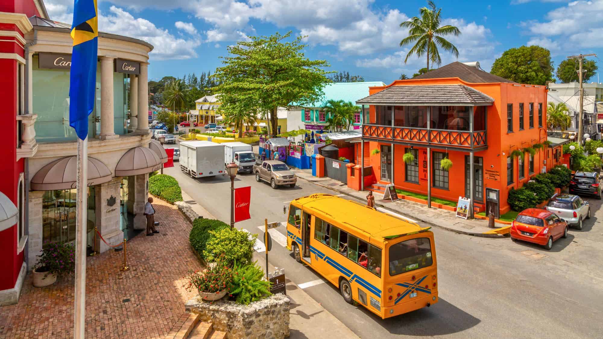 A lively street scene in Bridgetown features colorful buildings, local traffic, a bright yellow bus, and vibrant tropical storefronts.