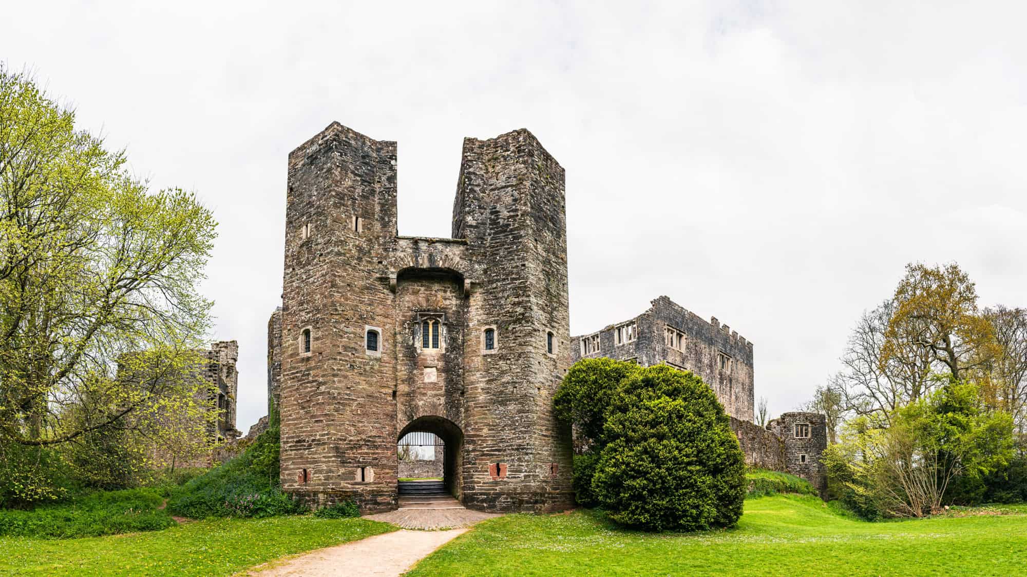 Large stone gatehouse of a partially ruined medieval castle stands amidst green lawns and trees, with the remaining castle walls visible behind.
