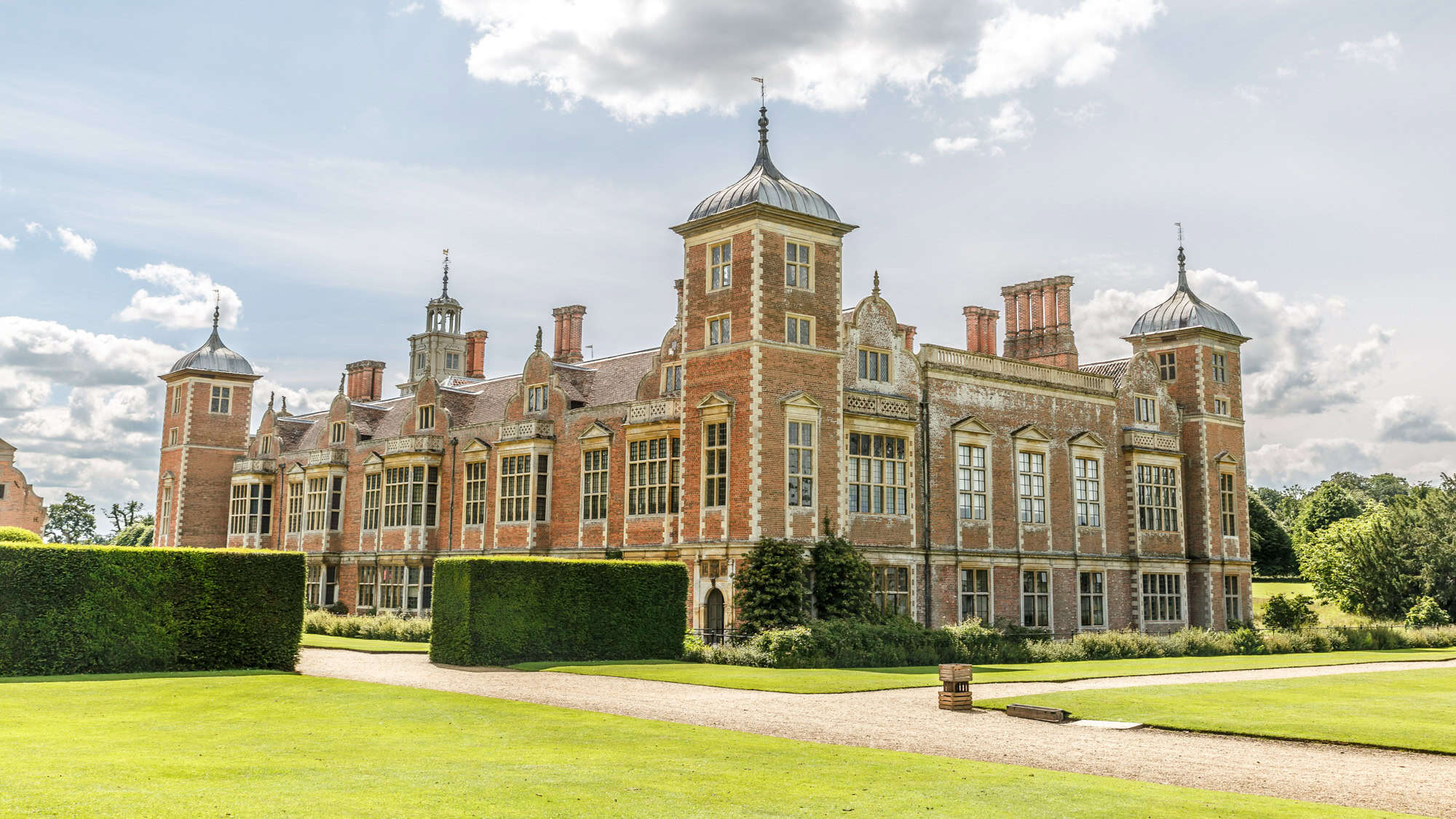 Grand Jacobean-style red brick mansion with elaborate towers and tall windows, set against neatly trimmed hedges and bright green lawns on a partly cloudy day.