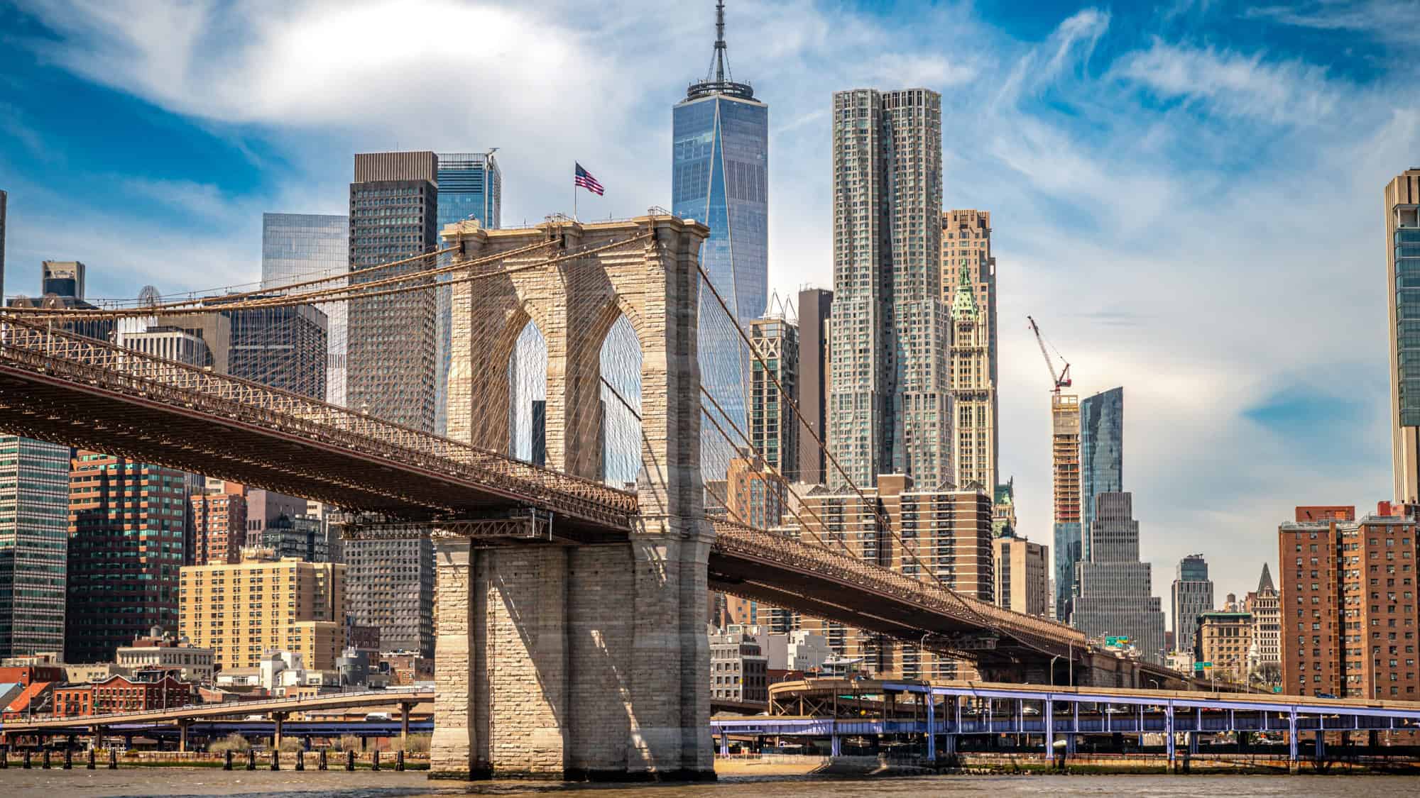 The Brooklyn Bridge stretches across the East River beneath a bright blue sky, with the towering Manhattan skyline and One World Trade Center rising in the background.