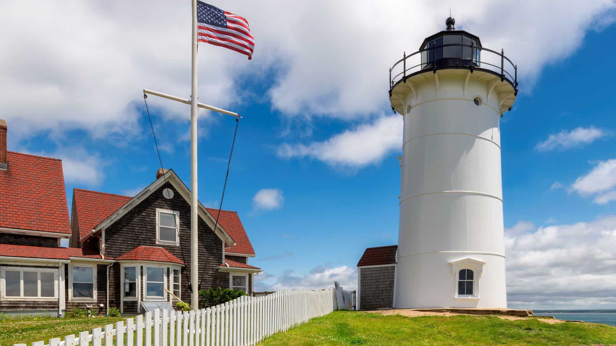 A tall, white lighthouse stands next to a traditional shingle-style keeper’s house with a red roof, an American flag flying proudly nearby under a blue sky.