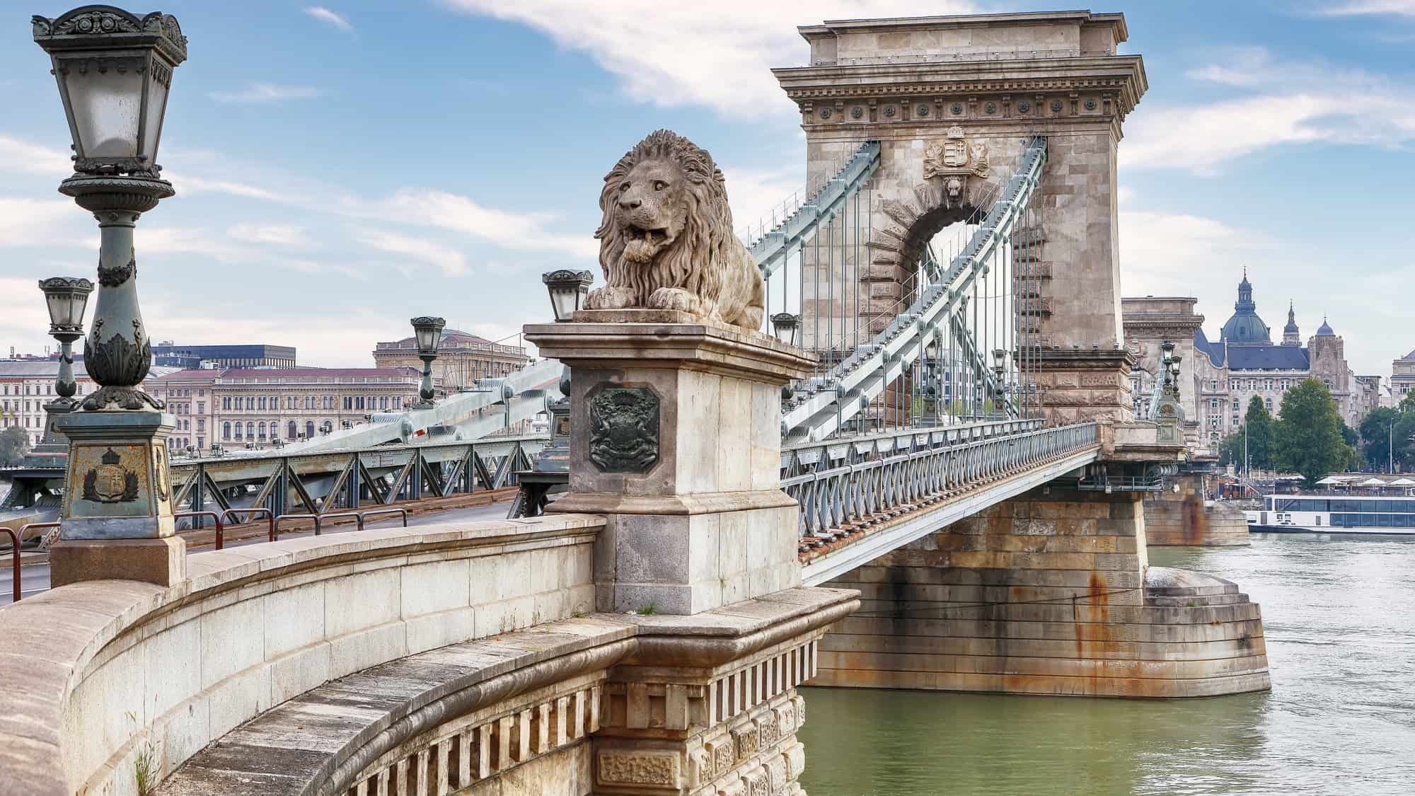 The historic Chain Bridge in Budapest features massive stone lions at its entrance and intricate ironwork, leading into the city over the Danube River.