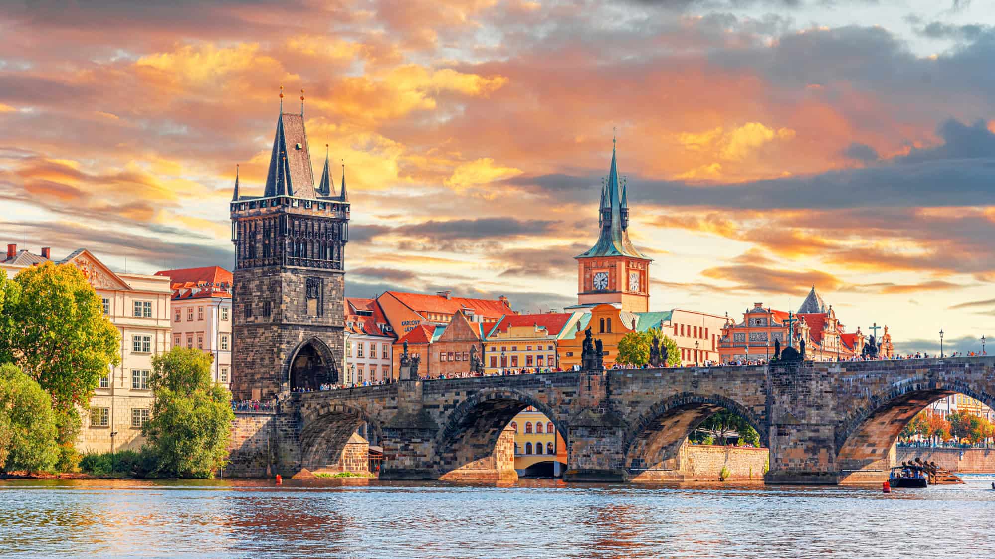 The Charles Bridge stretches across the Vltava River, with its Gothic towers and statues glowing under a vibrant, fiery sunset sky in Prague’s Old Town.