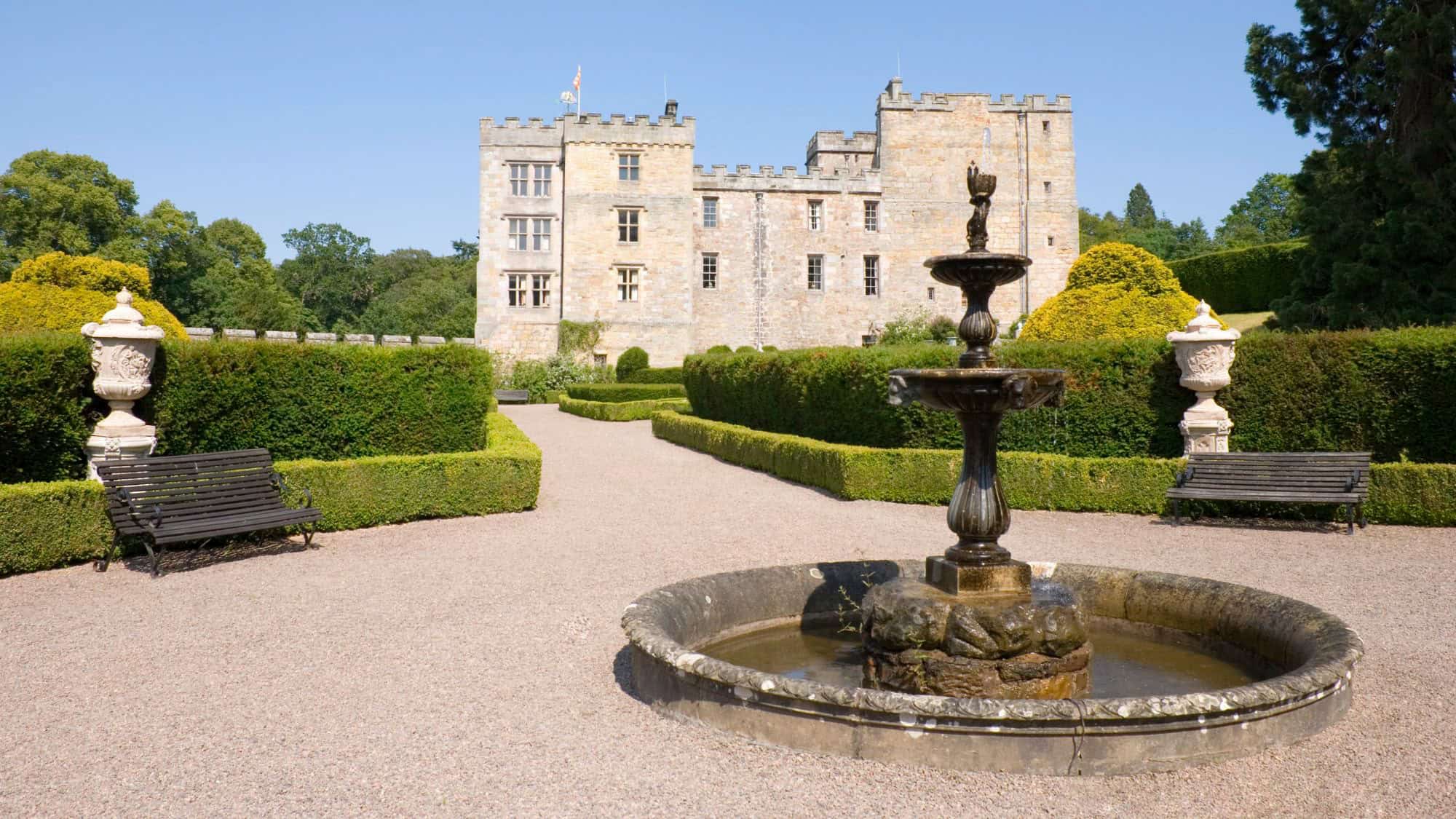 A stately stone castle with square towers overlooks a peaceful courtyard featuring formal hedges, benches, and a central tiered fountain.