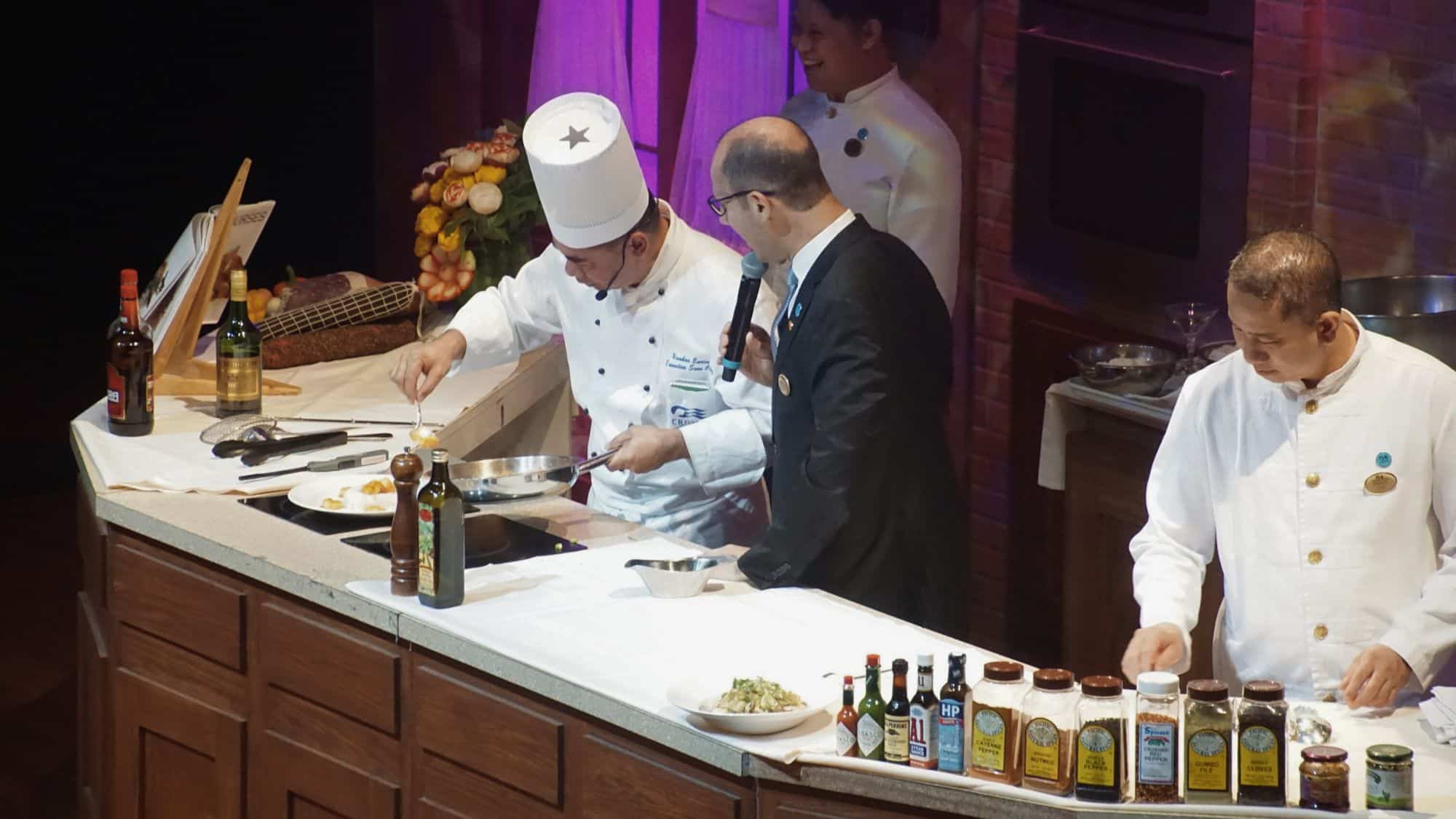 A chef in a white hat prepares food at a cooking station while being interviewed, surrounded by spices, sauces, and curious crew on a cruise ship stage.