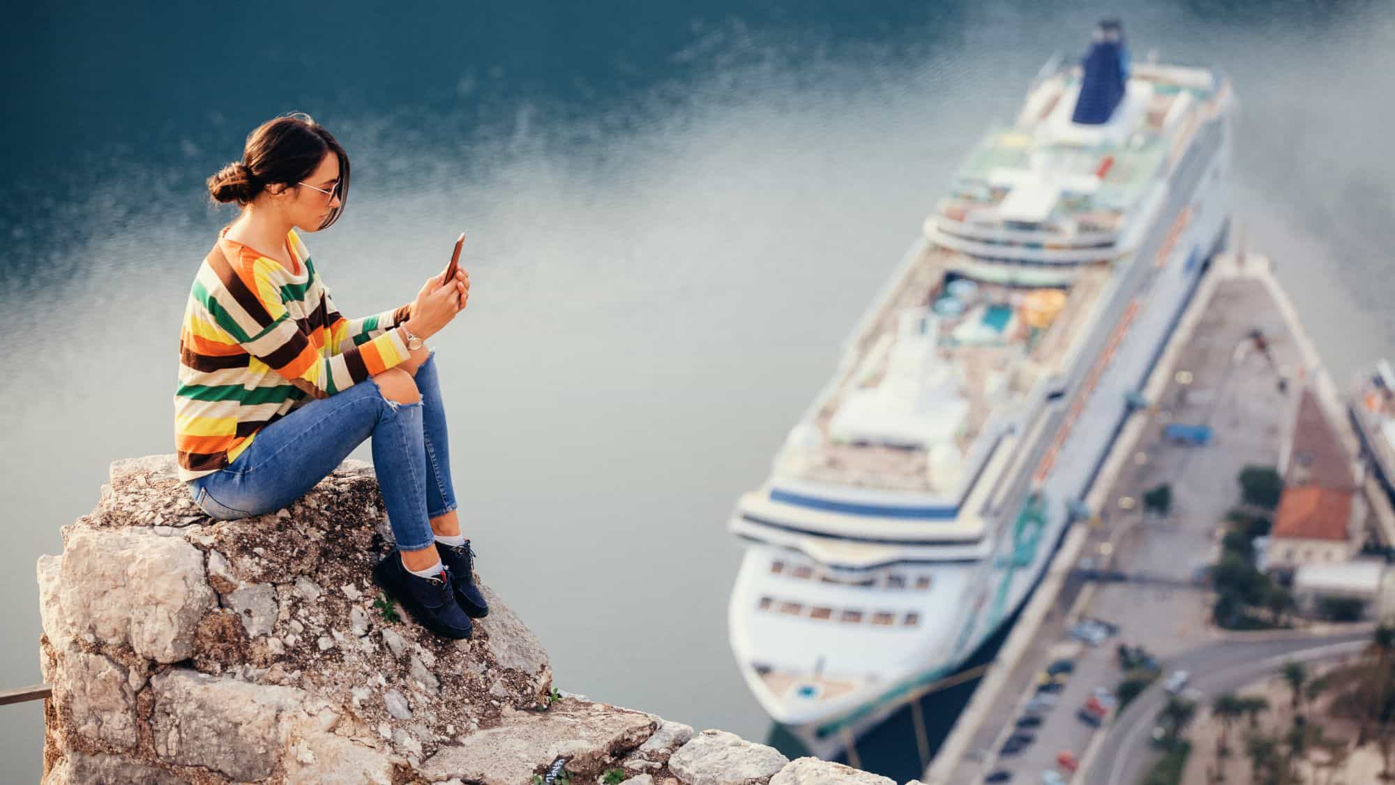 A woman in a striped sweater and jeans sits on a rocky cliff edge taking a selfie with a docked cruise ship far below in the water.