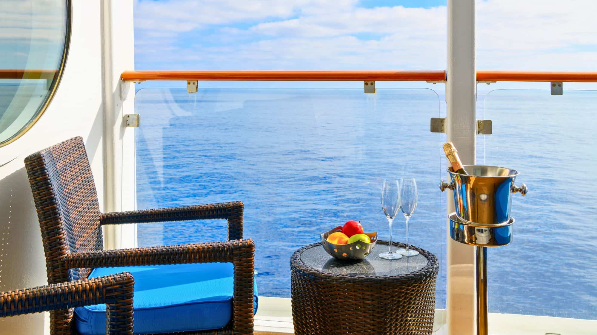 A cruise ship cabin balcony features wicker chairs, a fruit bowl, and a champagne bucket with two flutes, all overlooking the peaceful sea.