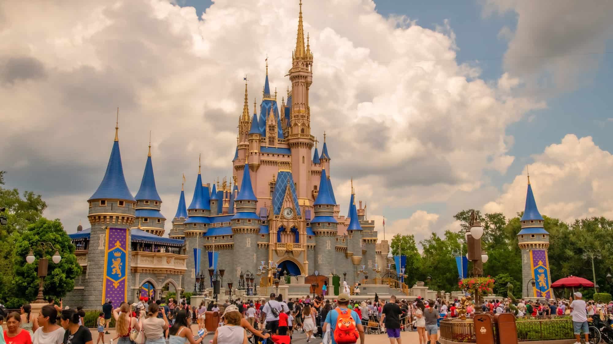 The iconic Cinderella Castle at Walt Disney World stands tall with blue and gold spires under a cloudy sky, as large crowds fill the surrounding plaza.