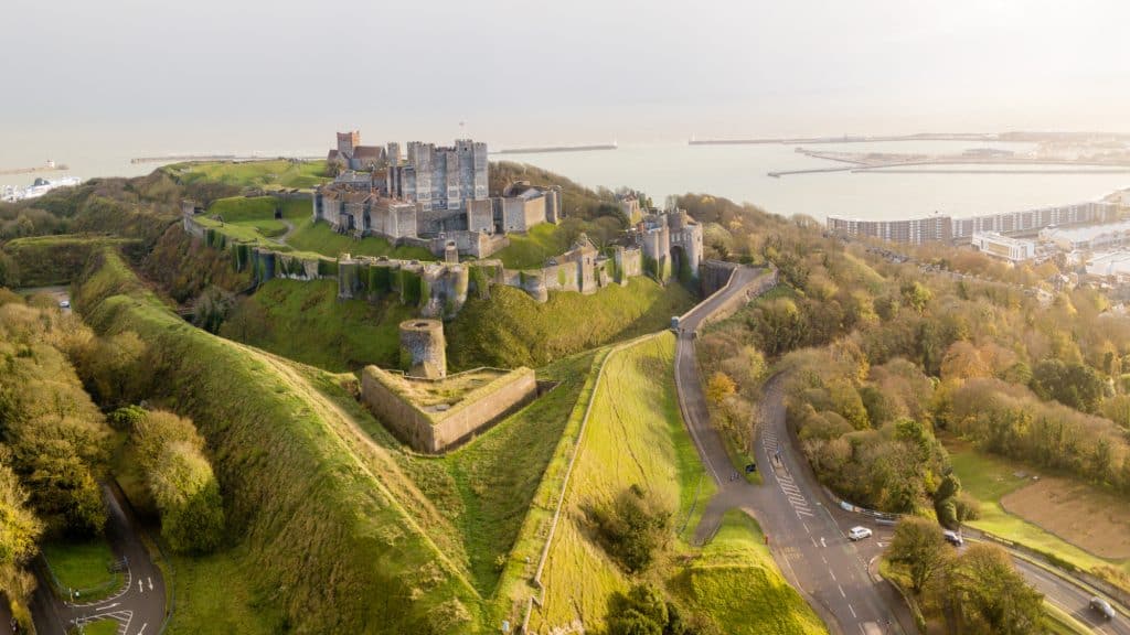 Expansive aerial view of Dover Castle and its layered defensive walls overlooking the English Channel, surrounded by rolling hills, trees, and the nearby port.
