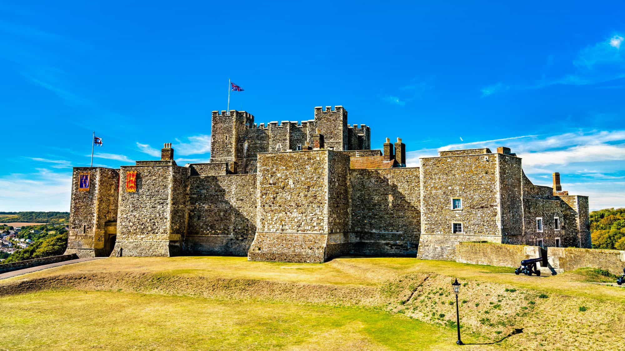 Massive stone walls and fortified towers of Dover Castle stand under a bright blue sky, with cannons on the grassy grounds and flags flying atop the battlements.
