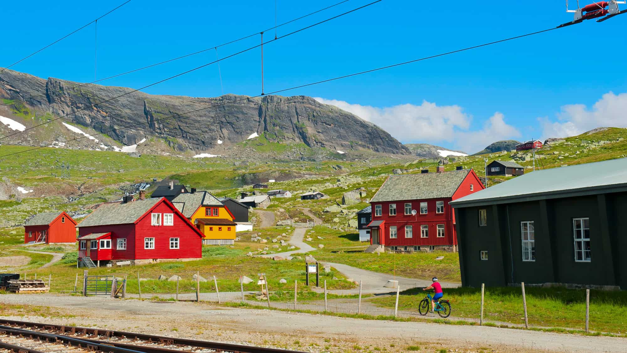 Red, yellow, and black wooden houses dot a quiet village nestled in the rocky, green Norwegian mountains, with a lone cyclist riding near railroad tracks under a bright blue sky.