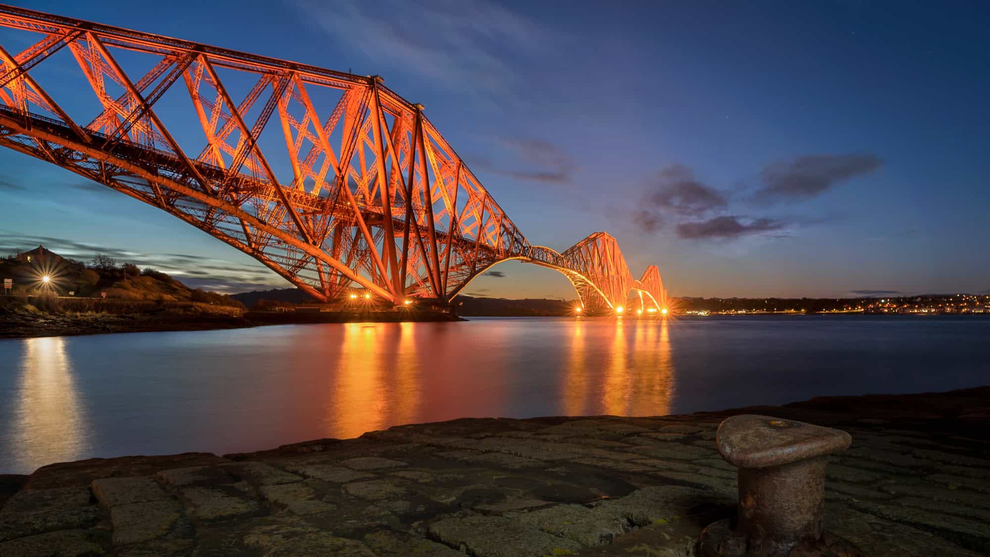 The iconic red cantilever Forth Bridge spans across the calm waters at twilight, its steelwork glowing with orange lights against a deep blue sky.