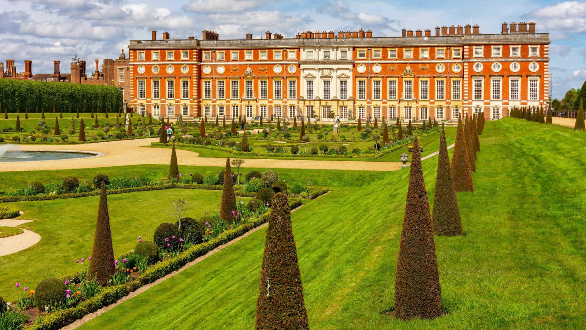 Elegant palace gardens in front of Hampton Court Palace with manicured lawns, cone-shaped topiary, blooming flowerbeds, and a grand baroque facade in warm red and white stone.