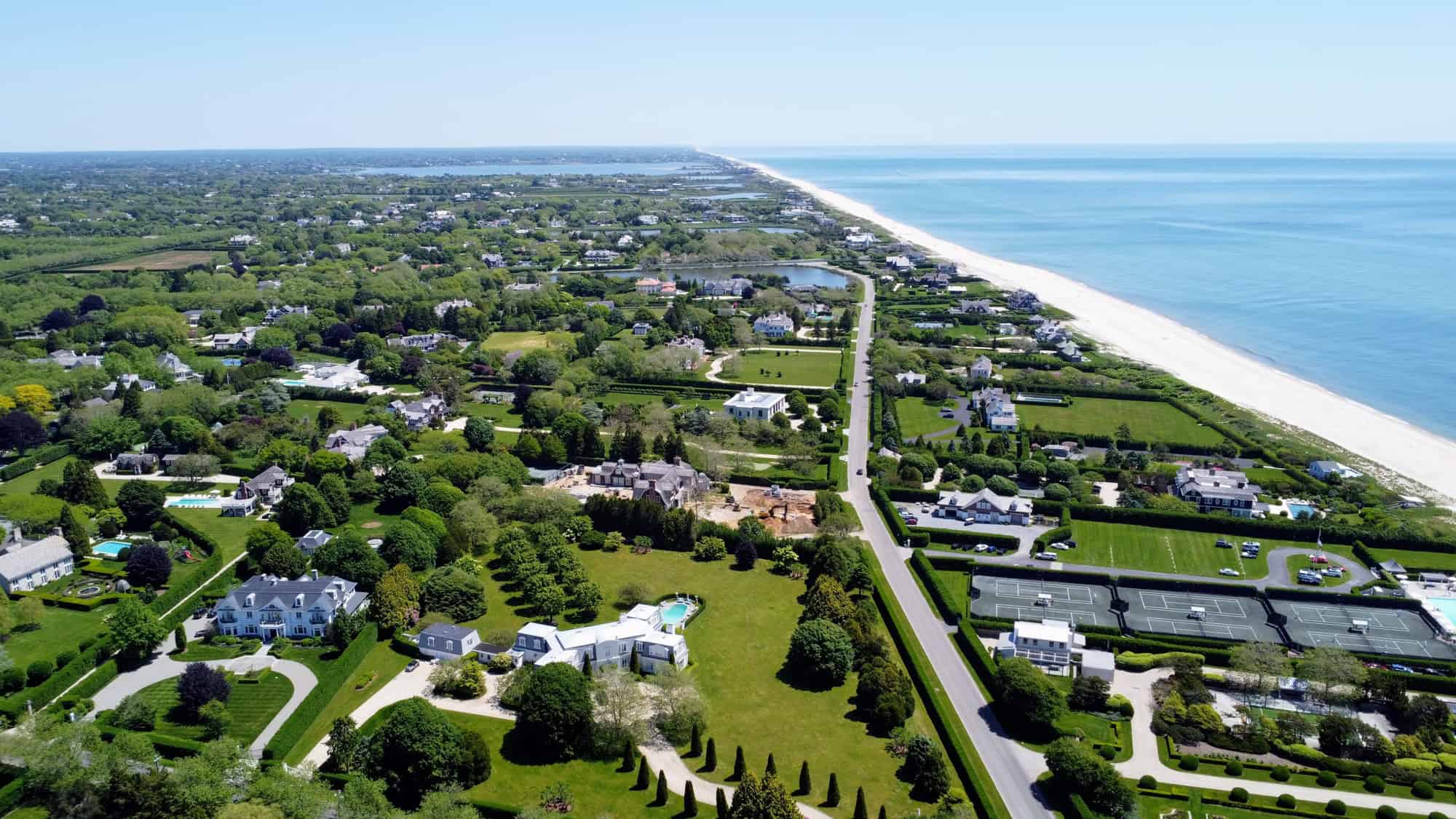 Lush green estates, tennis courts, and manicured lawns stretch along a road paralleling the white-sand beach and ocean on a sunny day.