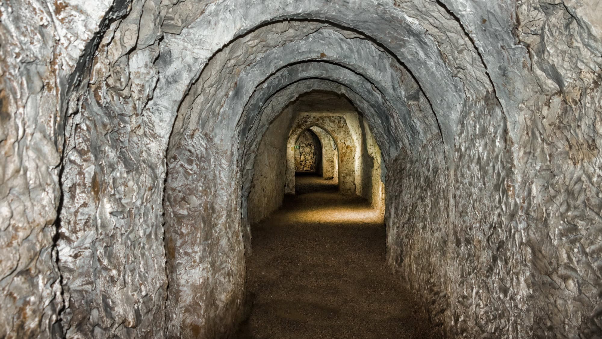 A narrow, arched stone tunnel with rough-hewn walls and gravel floor, lit intermittently by lamps and stretching into the distance in a castle or fort.