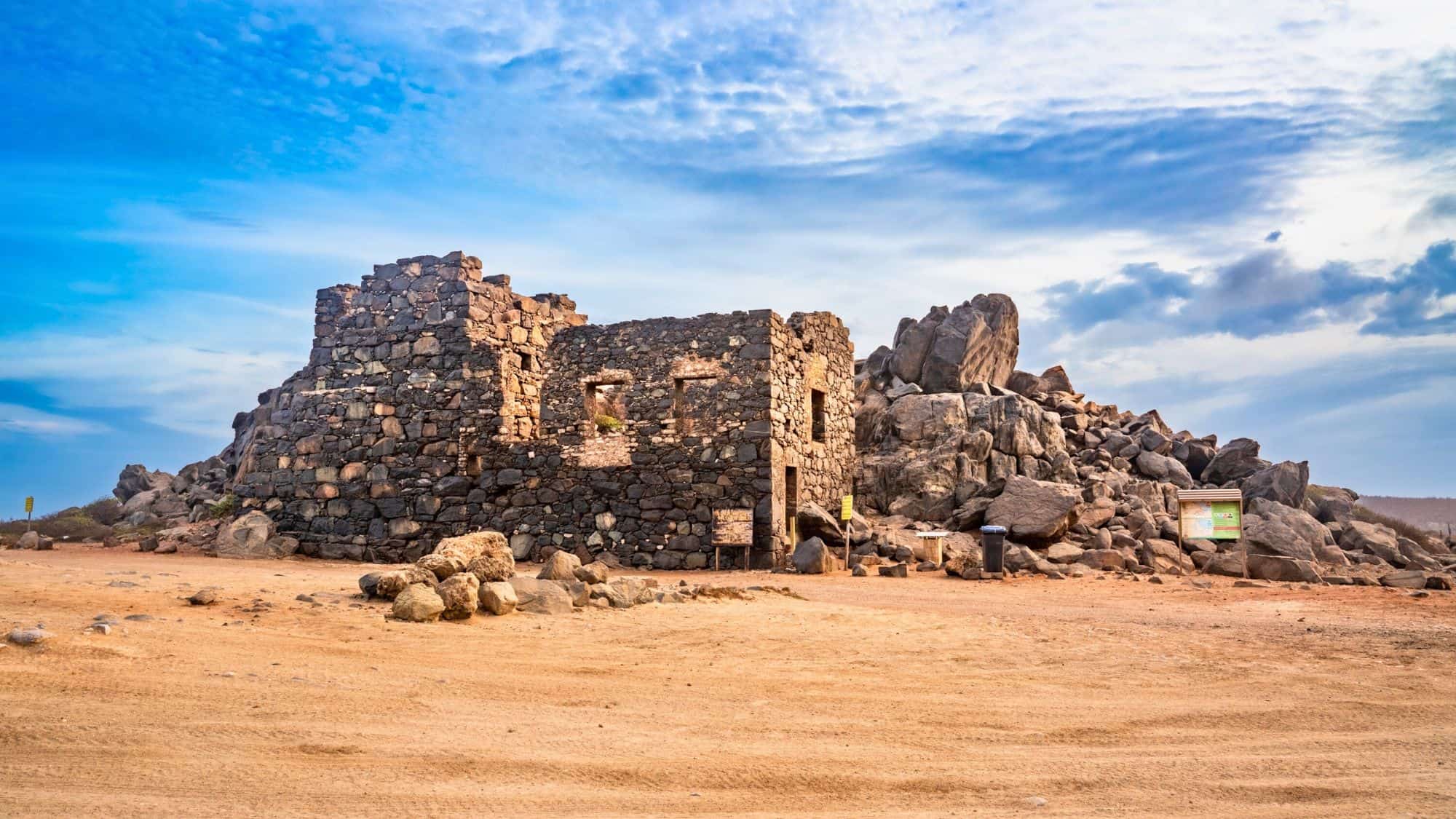 A historical stone structure set in a sandy, rocky environment. The ruins are accompanied by signage, suggesting a tourist or educational site.