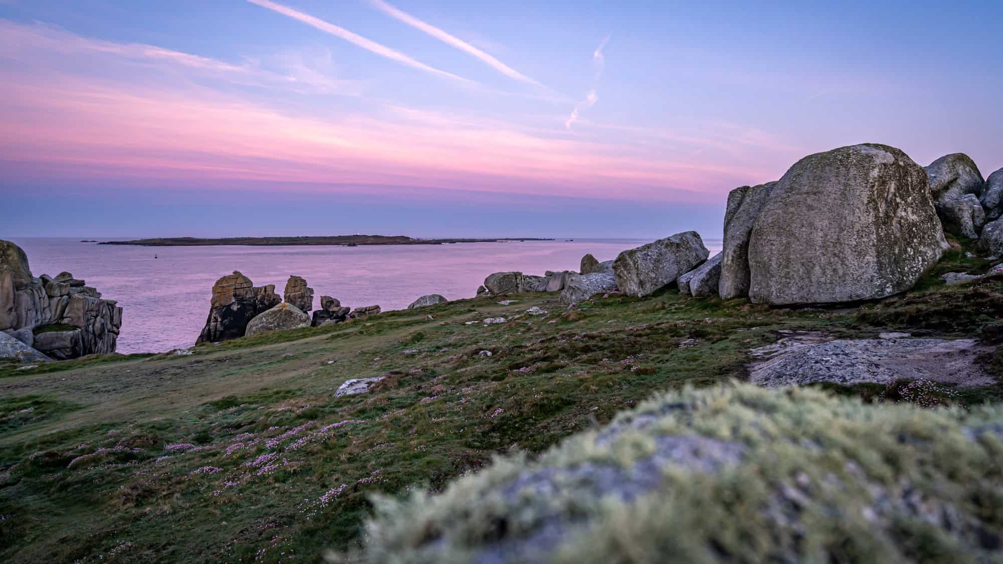 A serene coastal scene at sunset, with large granite boulders and grassy patches overlooking calm pink-tinged waters under a pastel sky.