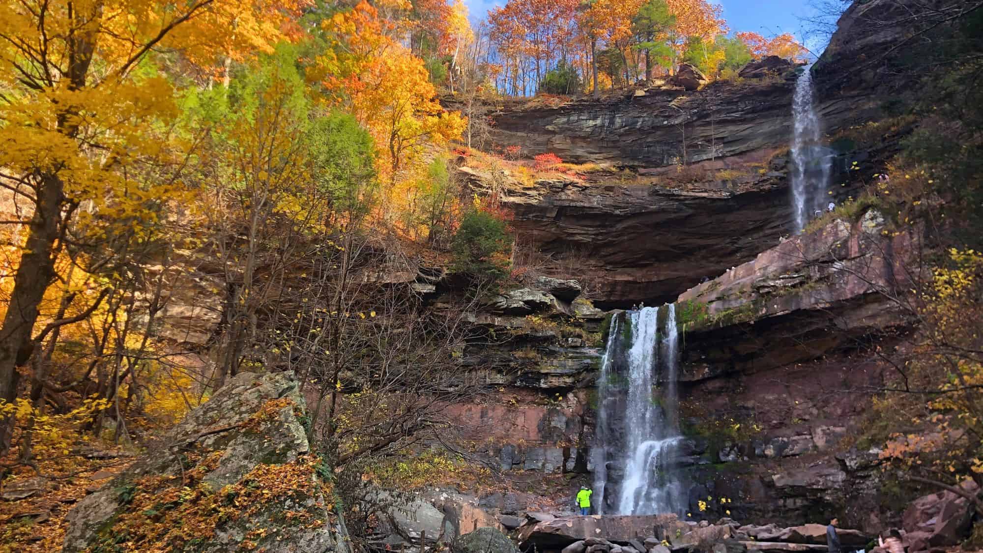 A two-tiered waterfall cascades down rugged rock cliffs framed by brilliant autumn foliage in shades of orange, yellow, and red, with hikers standing near the base for scale.