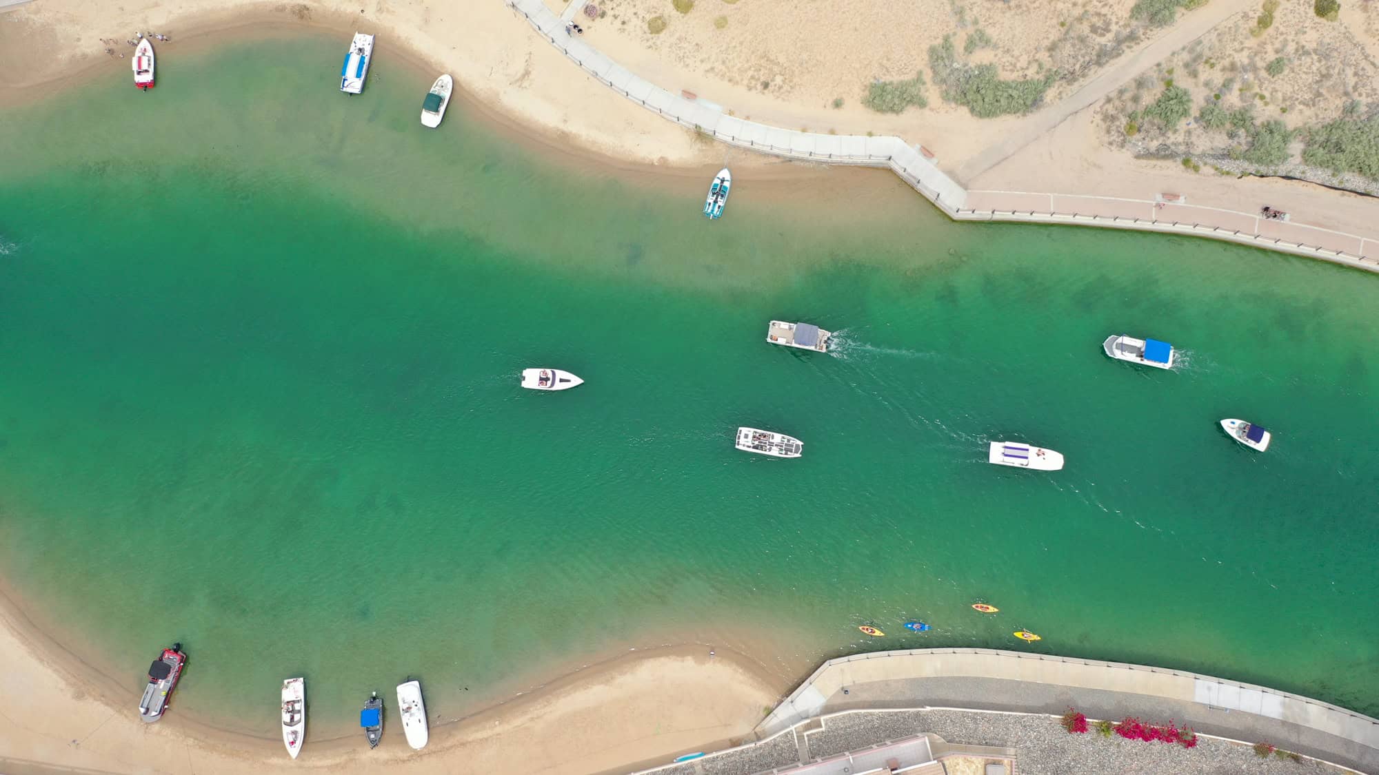 An aerial view shows boats cruising and anchored in a narrow, emerald-green waterway bordered by sandy shores and a curved pedestrian path.