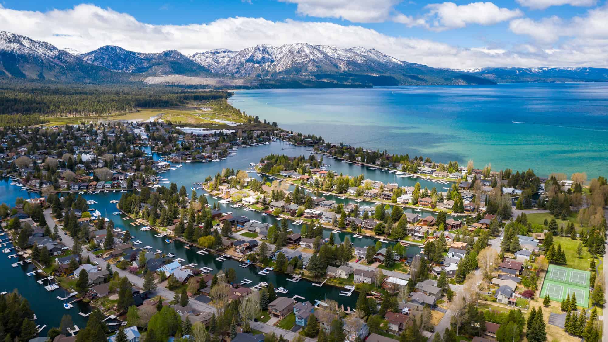 Aerial view of waterfront homes with private docks nestled among pine trees on Lake Tahoe, with snowcapped mountains and clear blue water in the distance.