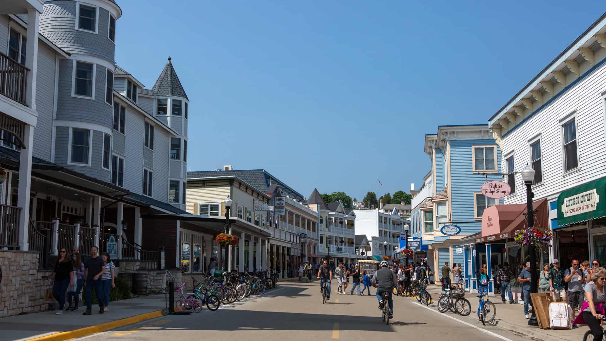A lively street scene on Michigan’s Mackinac Island, featuring Victorian-style buildings, pedestrians, and cyclists along a car-free road lined with shops and bike racks.