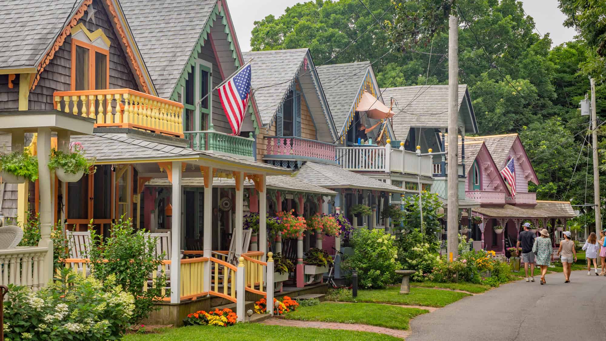 A charming row of brightly painted gingerbread cottages lines a quiet street, each with detailed trim, American flags, and blooming flowers.