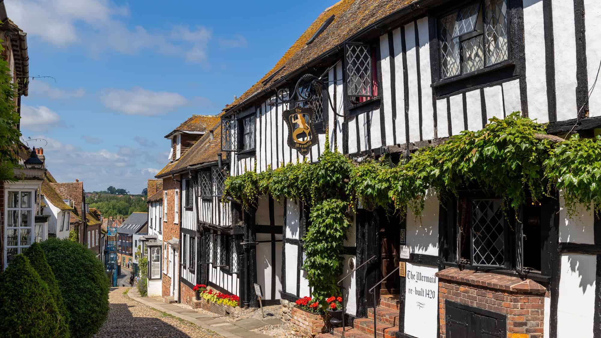 Tudor-style black-and-white timber-framed building with diamond-paned windows and ivy-covered frontage on a sloped cobblestone street, featuring a hanging sign reading "The Mermaid Inn, re-built 1420."