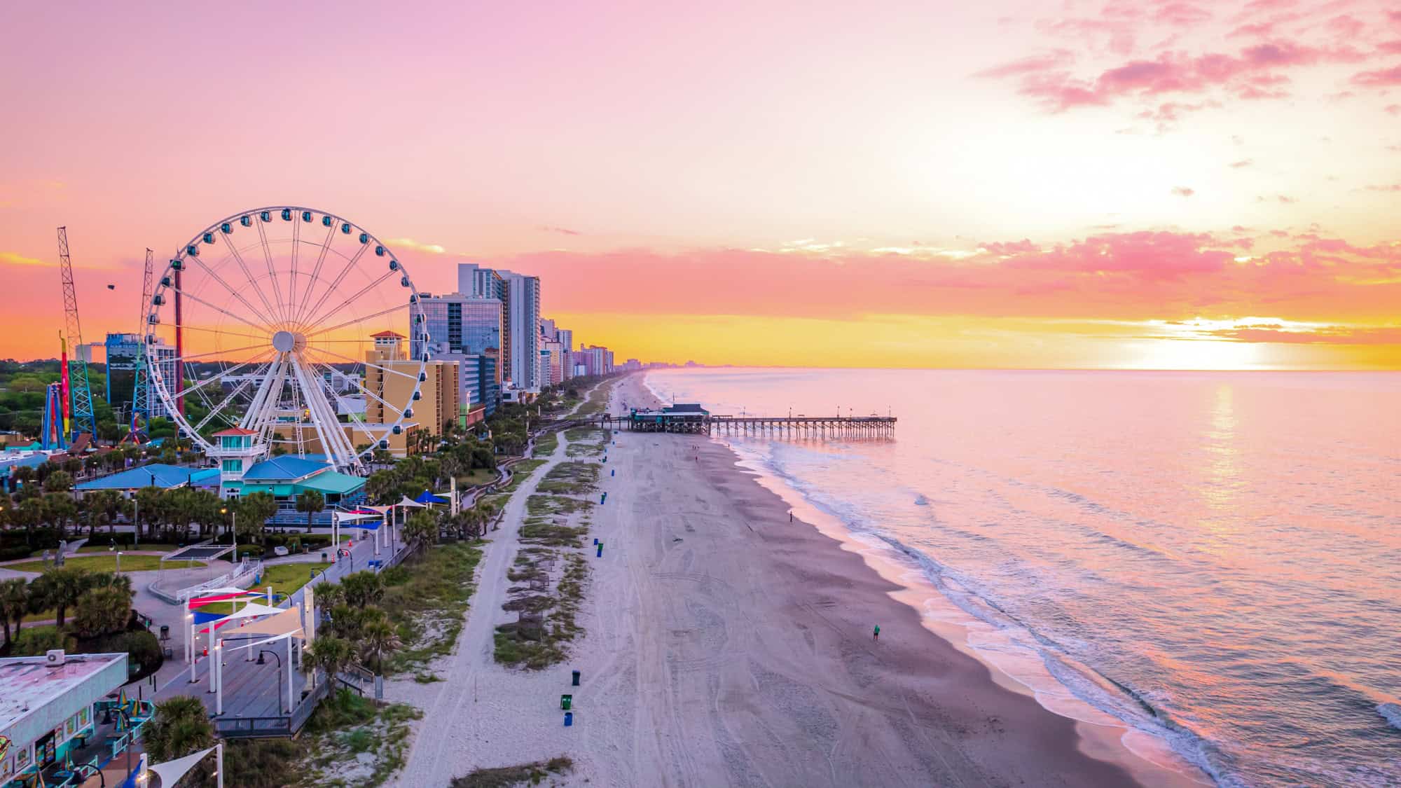 A pink and orange sunrise lights up the sky over Myrtle Beach, with the SkyWheel, high-rise hotels, and a pier visible along the shore.