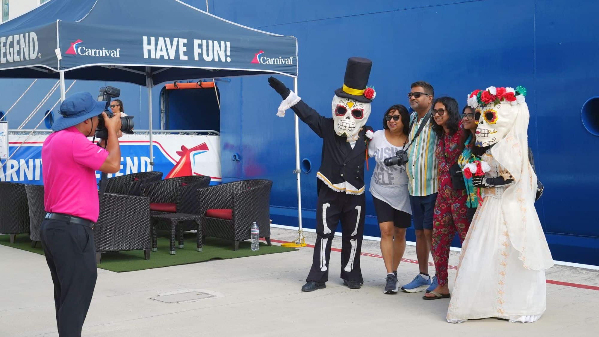 Passengers pose with costumed skeleton characters under a Carnival tent labeled “Have Fun!” while a photographer captures the moment on the dock.