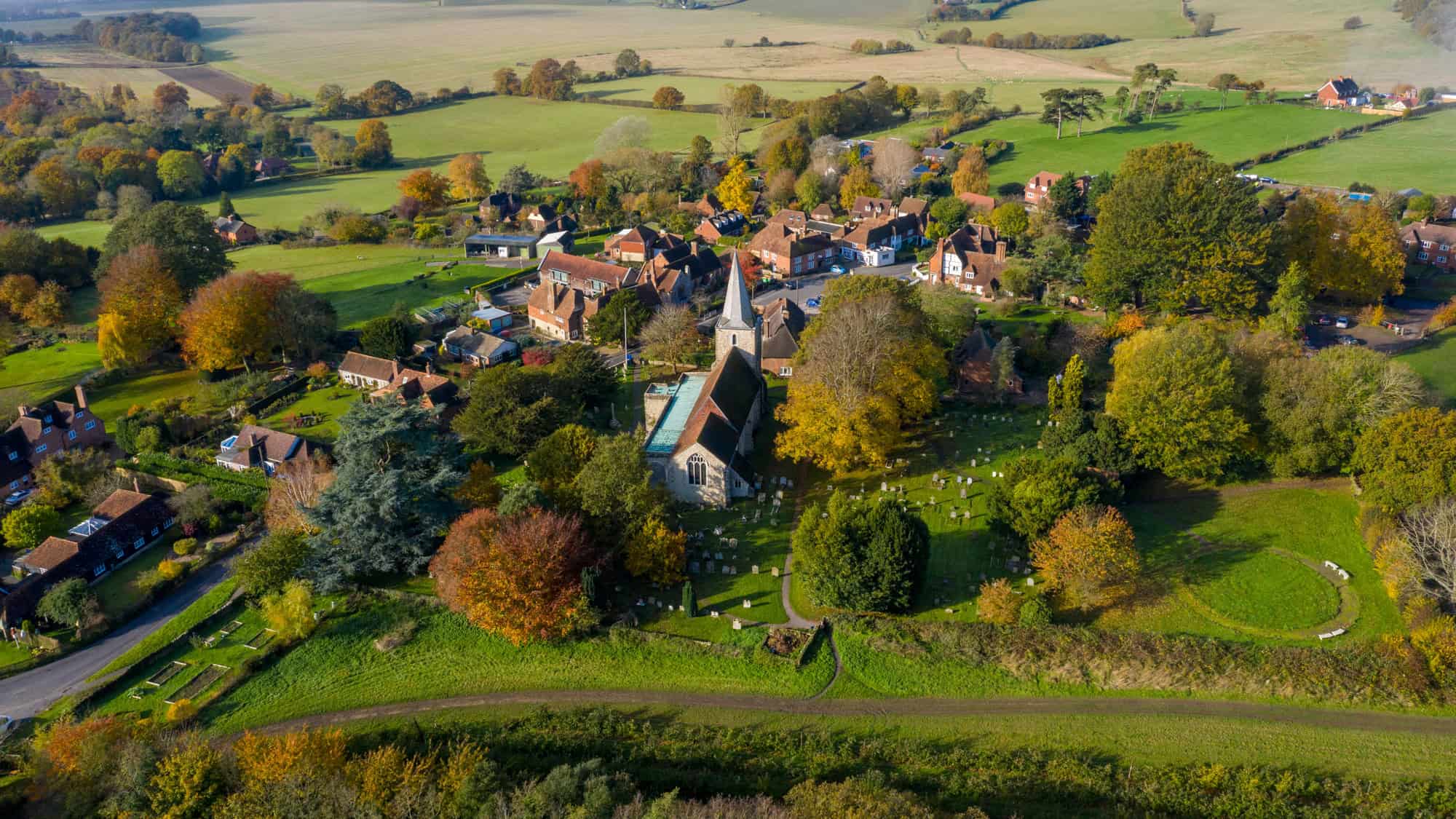 Aerial view of a quaint English village surrounded by rolling green fields in autumn, with a central stone church and gravestones nestled among colorful trees and charming brick homes.