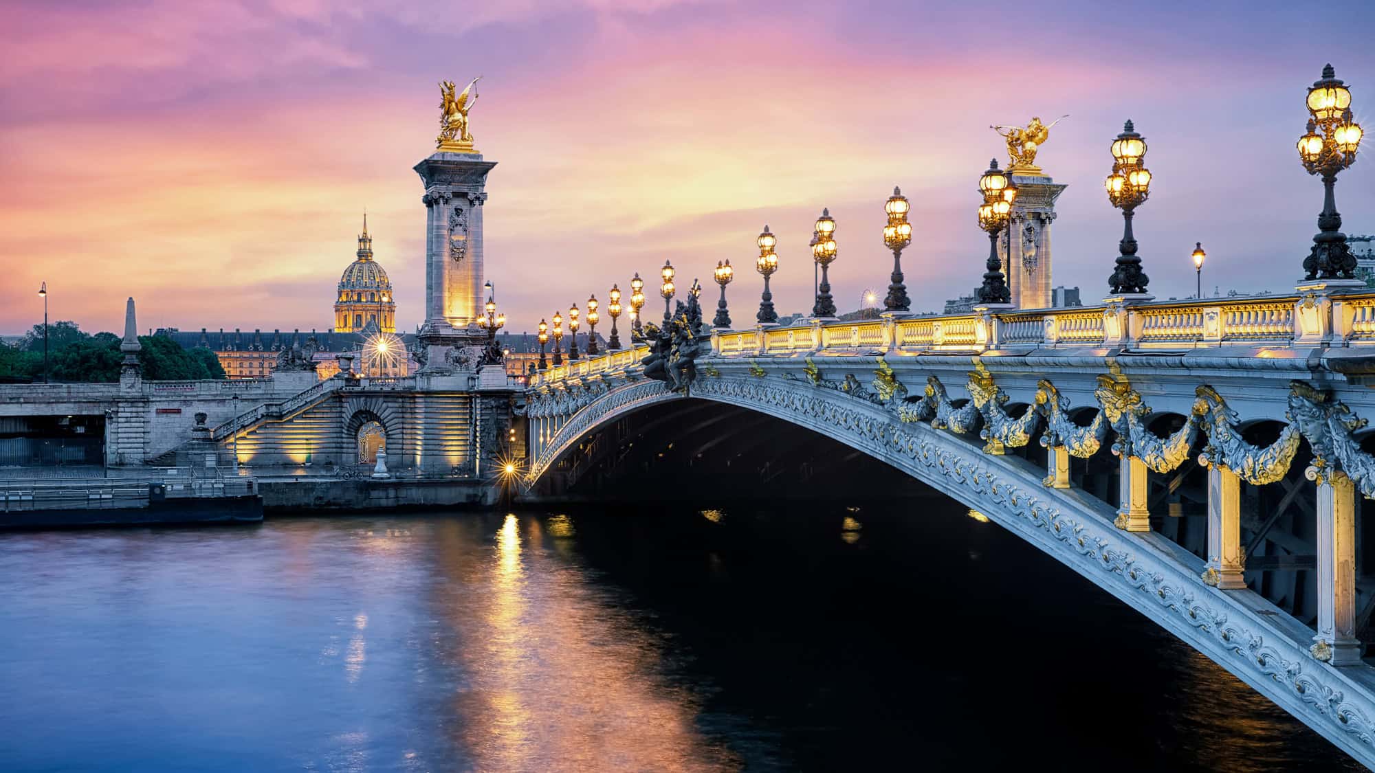 Paris’s ornate Pont Alexandre III bridge glistens at twilight with glowing streetlamps and gold statues, set against a pastel sunset sky and the golden dome of Les Invalides.