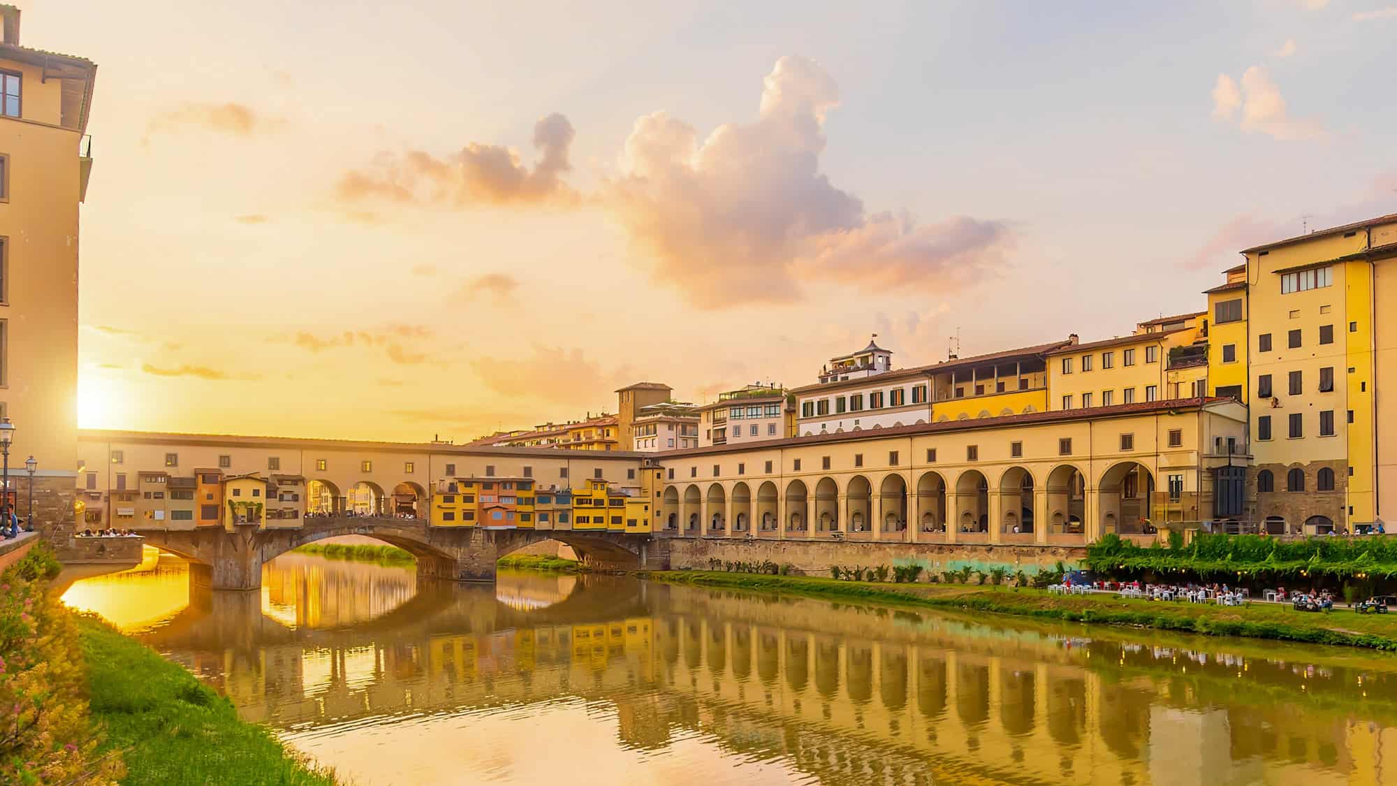 The Ponte Vecchio glows golden at sunset, with its colorful buildings and arches reflected in the calm waters of the Arno River, surrounded by historic architecture.