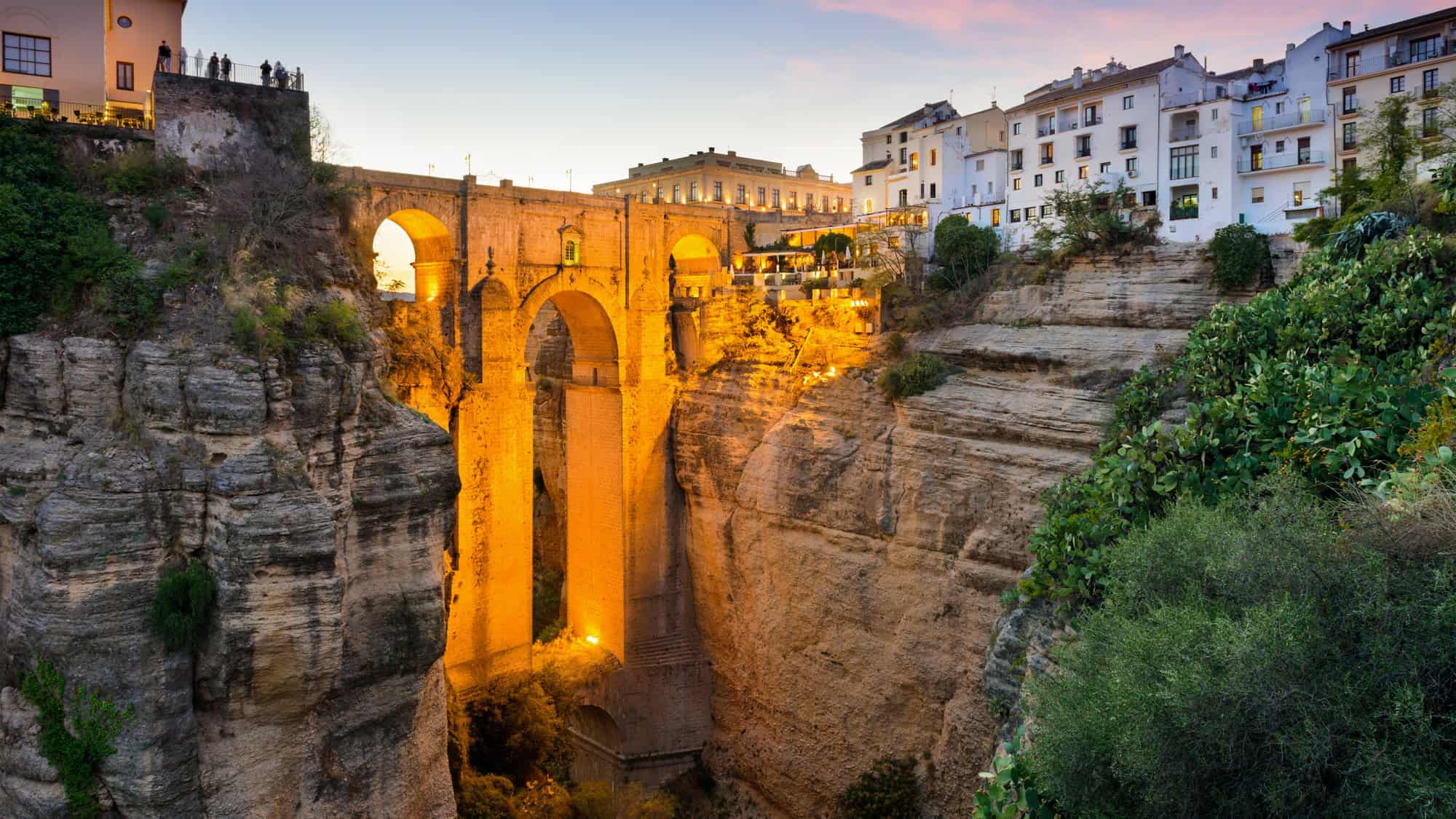 The Puente Nuevo bridge arches over a deep gorge in Ronda, lit warmly at dusk with whitewashed buildings perched dramatically on the cliffs above.
