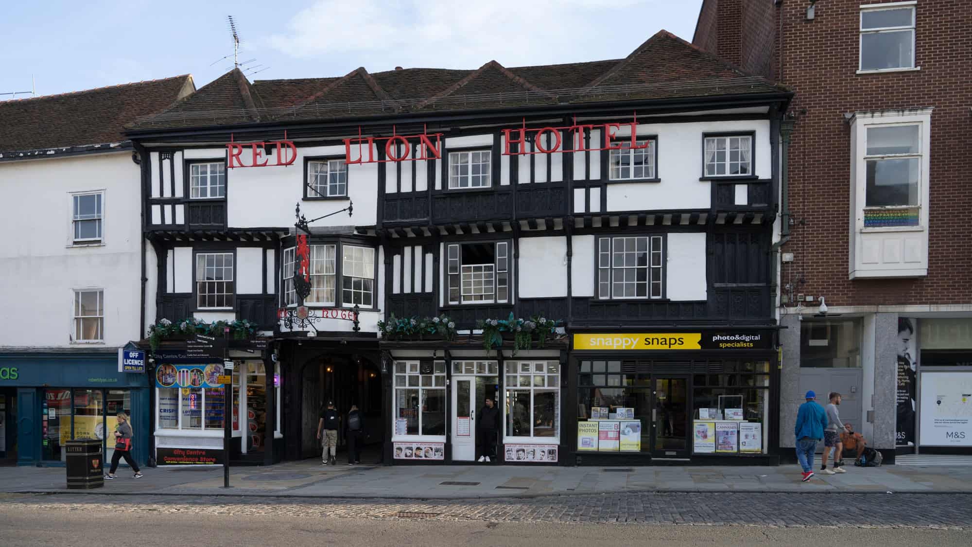 Historic timber-framed Red Lion Hotel with black-and-white exterior and red signage, flanked by modern shops on a bustling street in Salisbury.