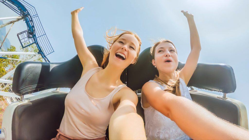 Two young women laugh and scream with arms raised while riding a roller coaster, capturing the thrill and energy of the moment against a bright blue sky. Two young women laugh and scream with arms raised while riding a roller coaster, capturing the thrill and energy of the moment against a bright blue sky.