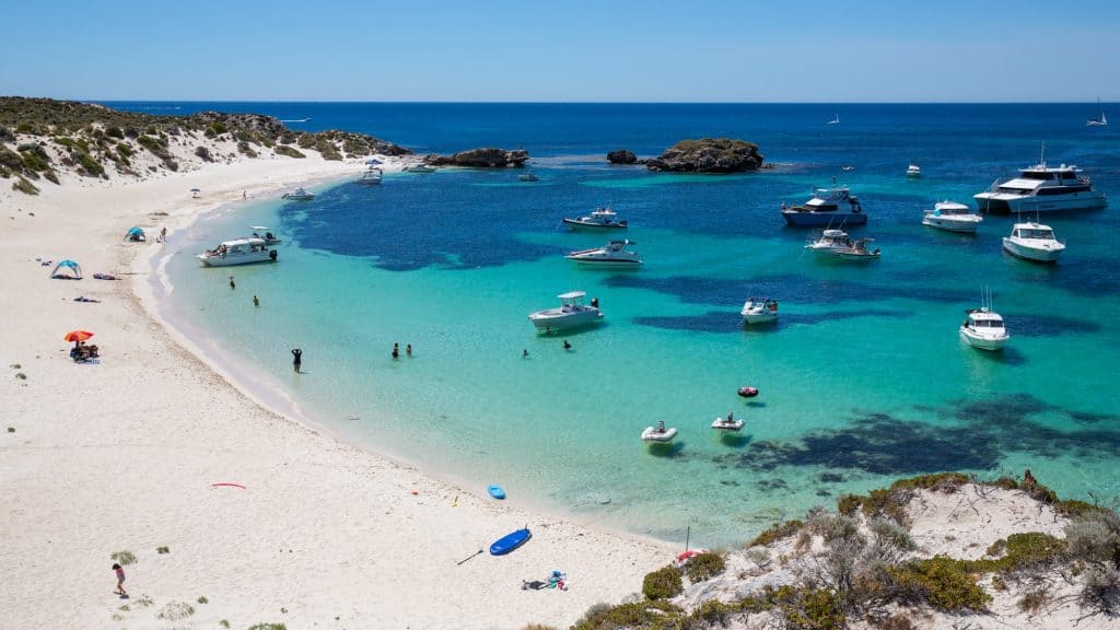 Dozens of boats float on the turquoise waters of a secluded sandy cove, with beachgoers lounging on the white shore and swimming in the clear sea under a sunny blue sky.