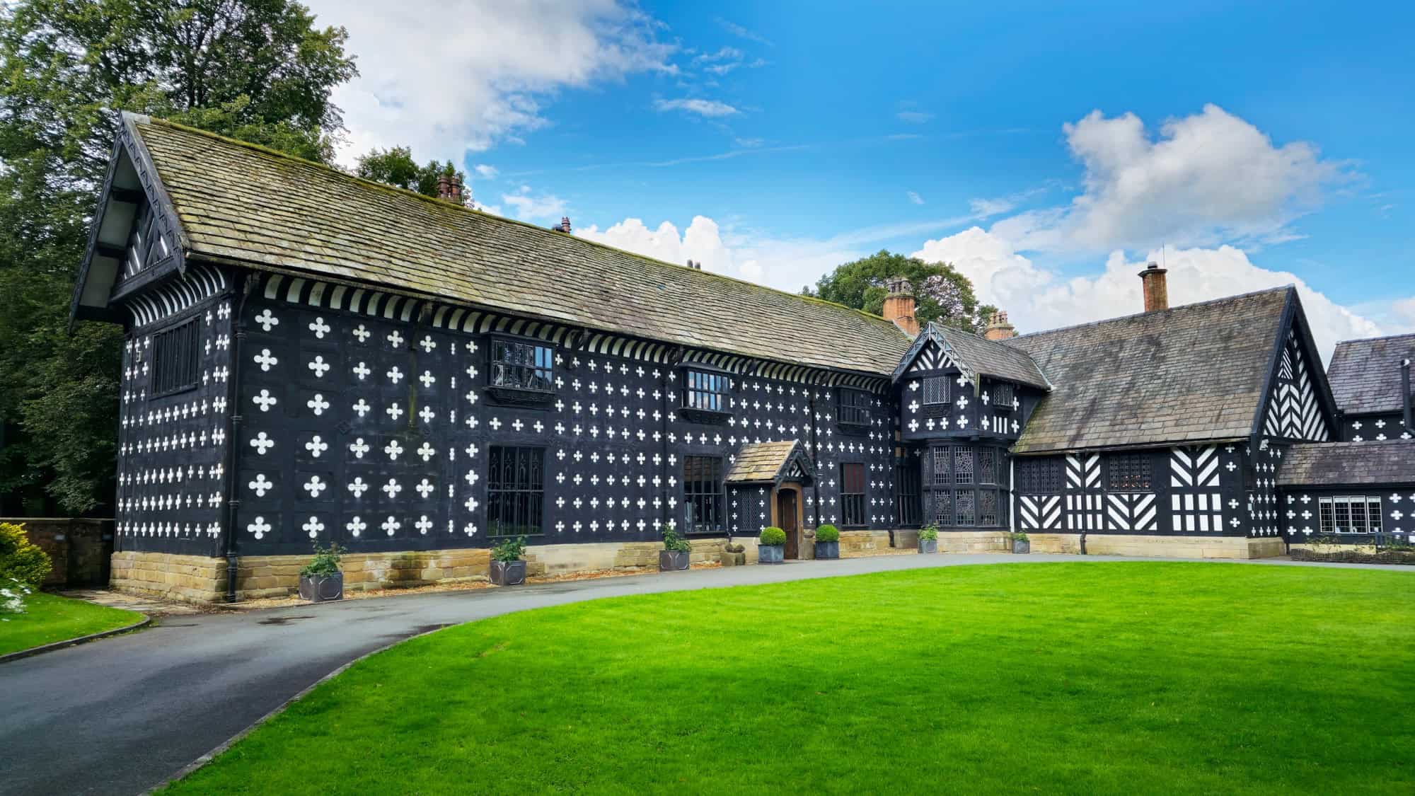 A striking black-and-white timber-framed Tudor manor with decorative quatrefoil patterns across the facade, set against a green lawn and surrounded by trees.